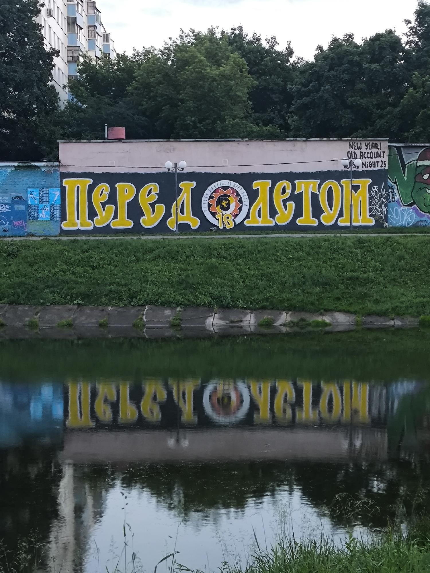The image shows a graffiti-covered wall adjacent to a body of water, likely a canal or river. The wall features large, yellow Cyrillic lettering that appears to read "ПЕРЕД ЛЕТОМ" (Before Summer). To the right of the lettering, there is graffiti of a green, cartoonish character, possibly a ninja turtle, and further to the right, some abstract graffiti. Above the yellow lettering, there are two light fixtures and some smaller white text that reads "NEW YEAR! OLD ACCOUNT NIGHT 25". A building, partially obscured by trees, is visible in the background. The foreground shows the reflection of the wall and its graffiti in the dark, rippling water, with some grass growing along the embankment. The scene appears to be in an urban environment, with graffiti and a waterfront.