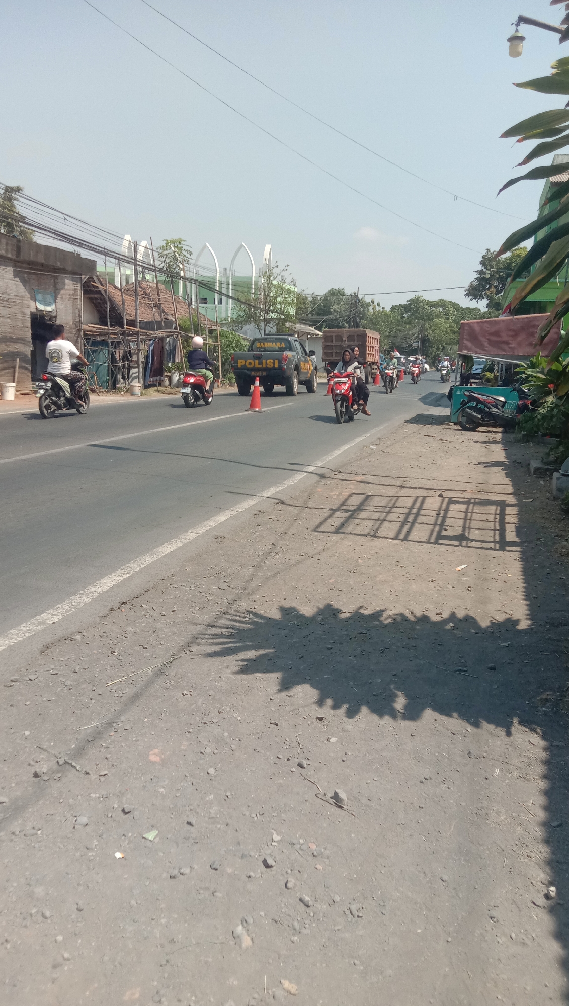 A sunny, hazy day in Karang Anyar, Indonesia, features a two-lane road with a single white line dividing the lanes. On the left side of the road, a man in a white shirt rides a motorcycle. Adjacent to the road is a low-lying structure with corrugated metal roofing. Further back on the left, a building with arched white elements and a green roof is visible, possibly a place of worship. On the right side of the road, a police truck with "POLISI" written on it is parked, with an orange traffic cone nearby. Several other motorcycles are visible on the road, some carrying passengers, heading in the same direction as the police truck. Lush green trees and foliage border the right side of the road. The road surface is asphalt and gravel. Shadows from trees and a structure are cast across the road and the ground on the right.