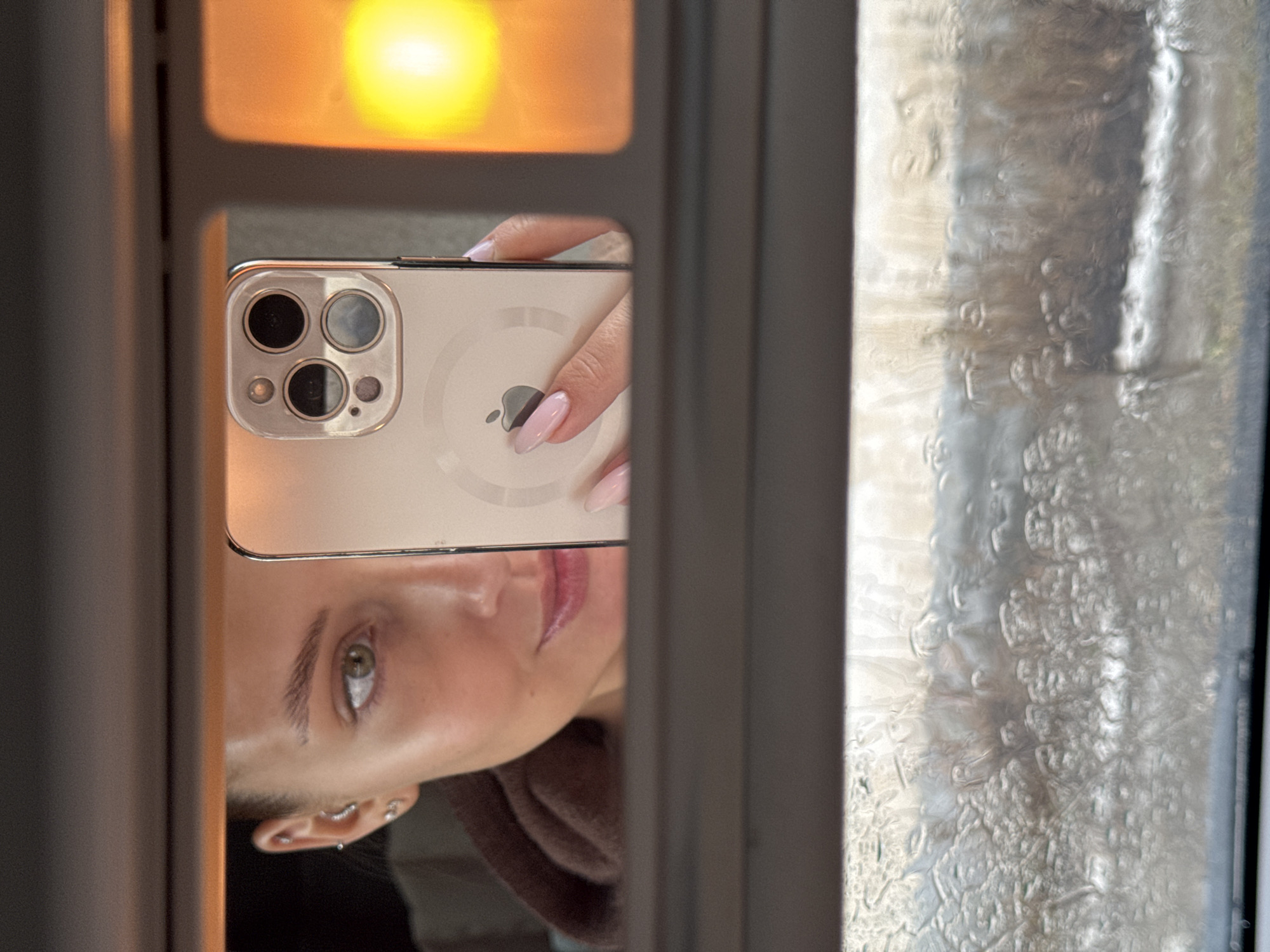 A person is reflected in a window pane, holding a light-colored smartphone with multiple camera lenses on the back. The person's face is partially visible, showing an eye and mouth. The phone's back is facing the viewer. Outside the window, there is a blurred, frosty or wet surface obscuring the view. The specific location in Chaikovsky, Russia, cannot be determined from the visual information provided. The environment appears to be indoors, based on the reflection and window.
