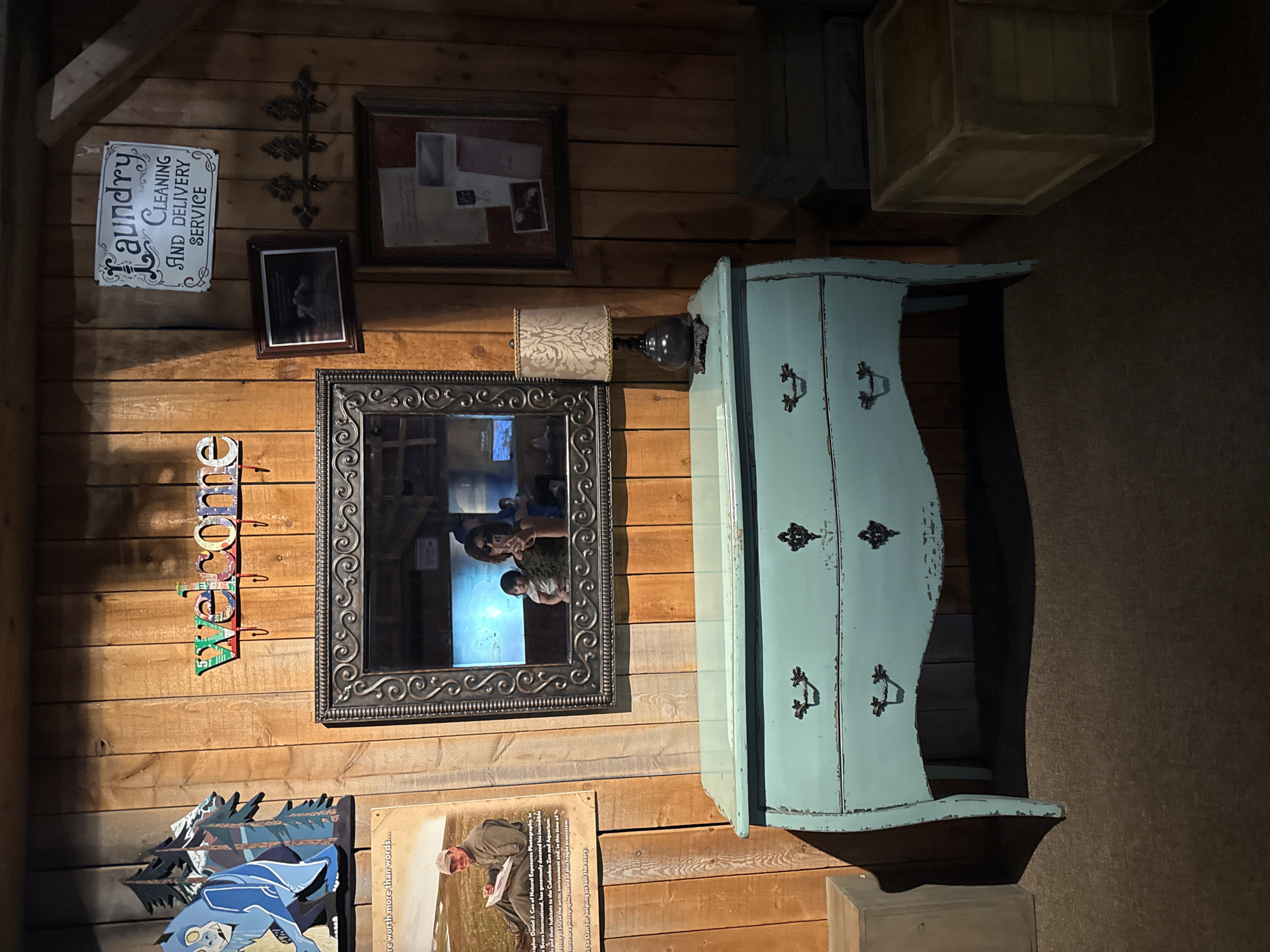 Here is a description of the image:
The image depicts a wooden wall adorned with various decorative items, presumably in an interior setting. Located in Powell, Ohio, the wall showcases a "Laundry Cleaning and Delivery Service" sign, a framed photo or document, a decorative metal piece resembling a stylized plant or branch, and a framed mirror reflecting people inside a building. Further down, the wall is decorated with a "Welcome" sign made of colorful letters, an image of blue birds and trees, and a framed photo or display board of natural exposures photography. To the right of the mirror stands a light-blue painted cabinet with drawers and black decorative drawer pulls. Above the cabinet are two wooden crates or boxes stacked on top of each other. The entire scene appears to be curated as part of an interior design or decorative arrangement.