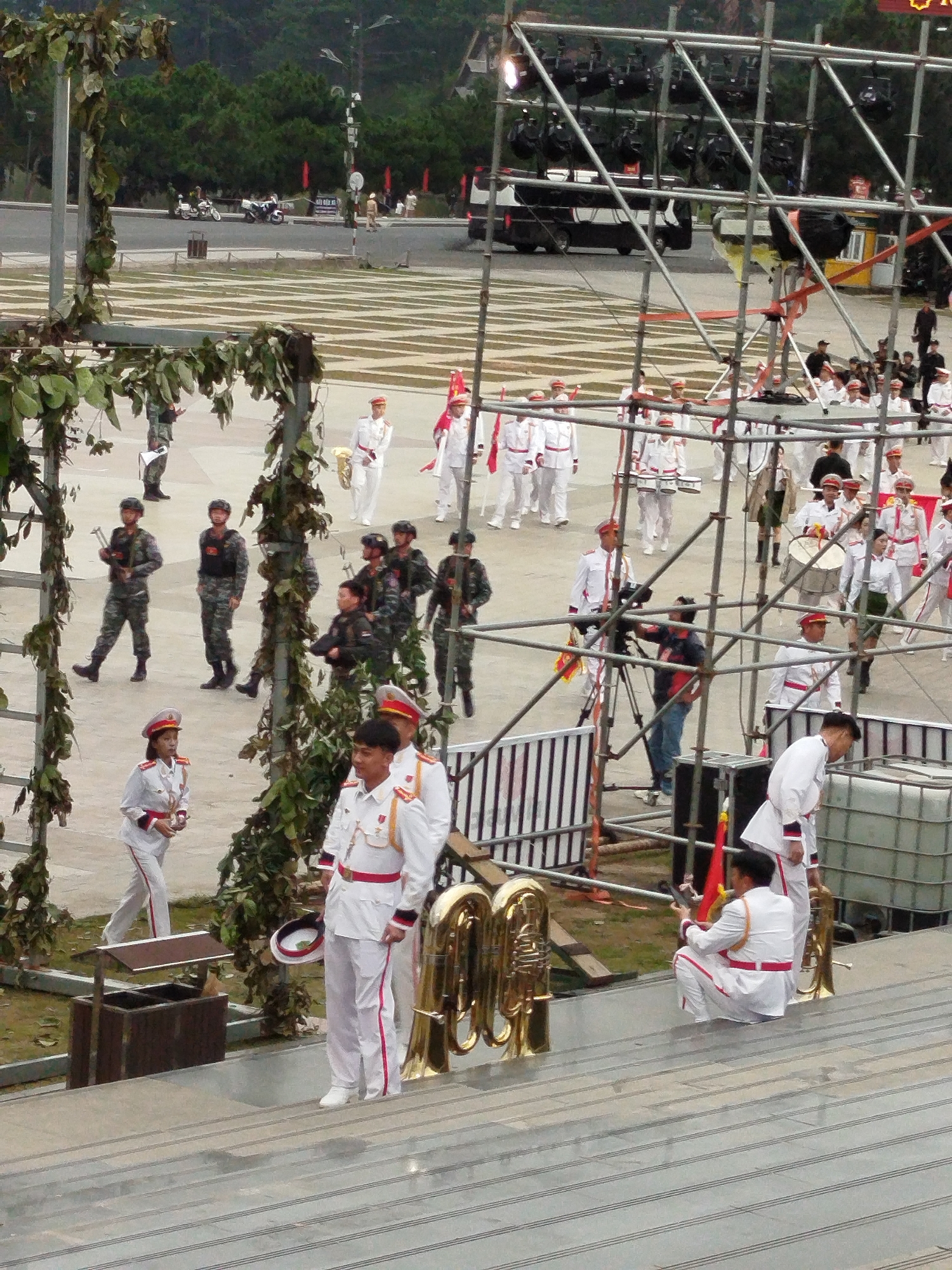 The image captures a scene of a formal parade or ceremony taking place outdoors, likely in Da Lat, Vietnam. A group of people in uniform are the primary subjects. Many are dressed in white ceremonial uniforms with red accents, suggesting a military or official capacity. Some individuals are holding musical instruments, including large brass tubas, indicating a band performance is part of the event. Others are in camouflage military uniforms, carrying what appear to be rifles.
The setting includes a large open paved area, possibly a plaza or parade ground, with a raised platform or grandstand in the foreground where the viewer is positioned. In the background, there are trees, a road with motorcycles, and a large bus. A scaffolding structure with lighting equipment is erected, suggesting a performance or broadcast is being staged. The weather appears overcast, and the time of day seems to be daytime, perhaps late afternoon, given the lighting.
One person in a white uniform is holding a ceremonial hat. Another person in a similar uniform is crouching down, holding what could be a camera or another piece of equipment. The overall mood seems formal and orderly, typical of such events. There are no discernible emotions overtly expressed by the subjects. No visible text is present in the image.