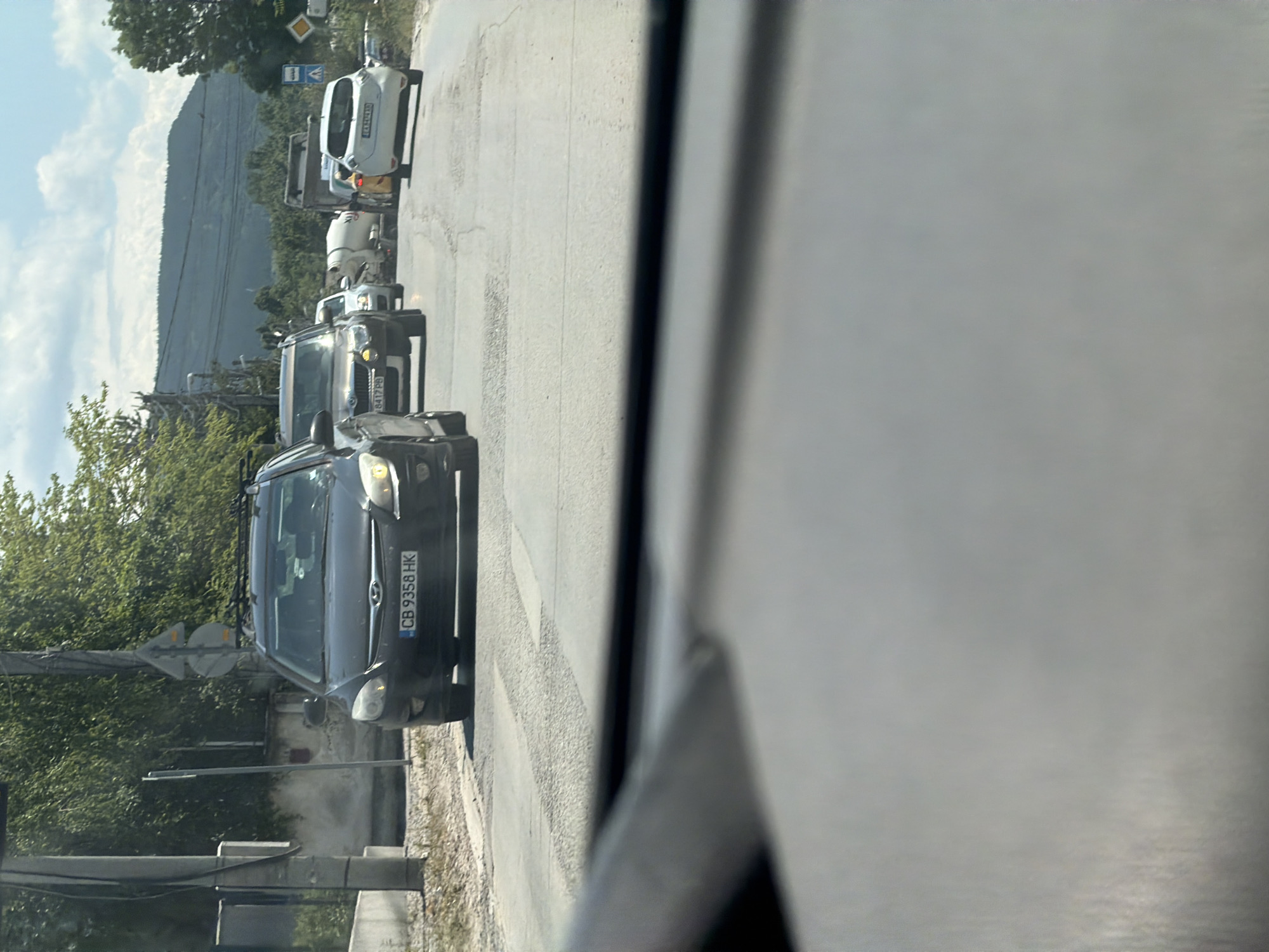 The image depicts a traffic jam on a two-lane road in a semi-urban or suburban area, likely in Sofia, Bulgaria. The foreground shows the interior of a vehicle, with the dashboard and windshield taking up the bottom portion of the frame. The road surface is asphalt and appears cracked in several sections.
In the middle ground, several vehicles are visible, suggesting slow-moving traffic or a complete standstill. The most prominent vehicle is a dark-colored hatchback with the license plate "CB 9358 HK". To its right, a silver SUV and a white passenger car are also present. Further down the road, a white cement mixer truck is visible, along with other vehicles, including a white electric vehicle with the license plate "EA 8457 AE". The presence of a cement mixer truck might indicate construction activity nearby.
The background features a line of trees and hills under a partly cloudy sky. There are also utility poles and wires along the side of the road. Road signs are visible, including a blue rectangular sign with a white symbol and a yellow diamond-shaped sign. The overall impression is a common scene of traffic congestion, possibly during daylight hours. The architectural elements, vegetation, and the style of vehicles and license plates are consistent with a location in Bulgaria.