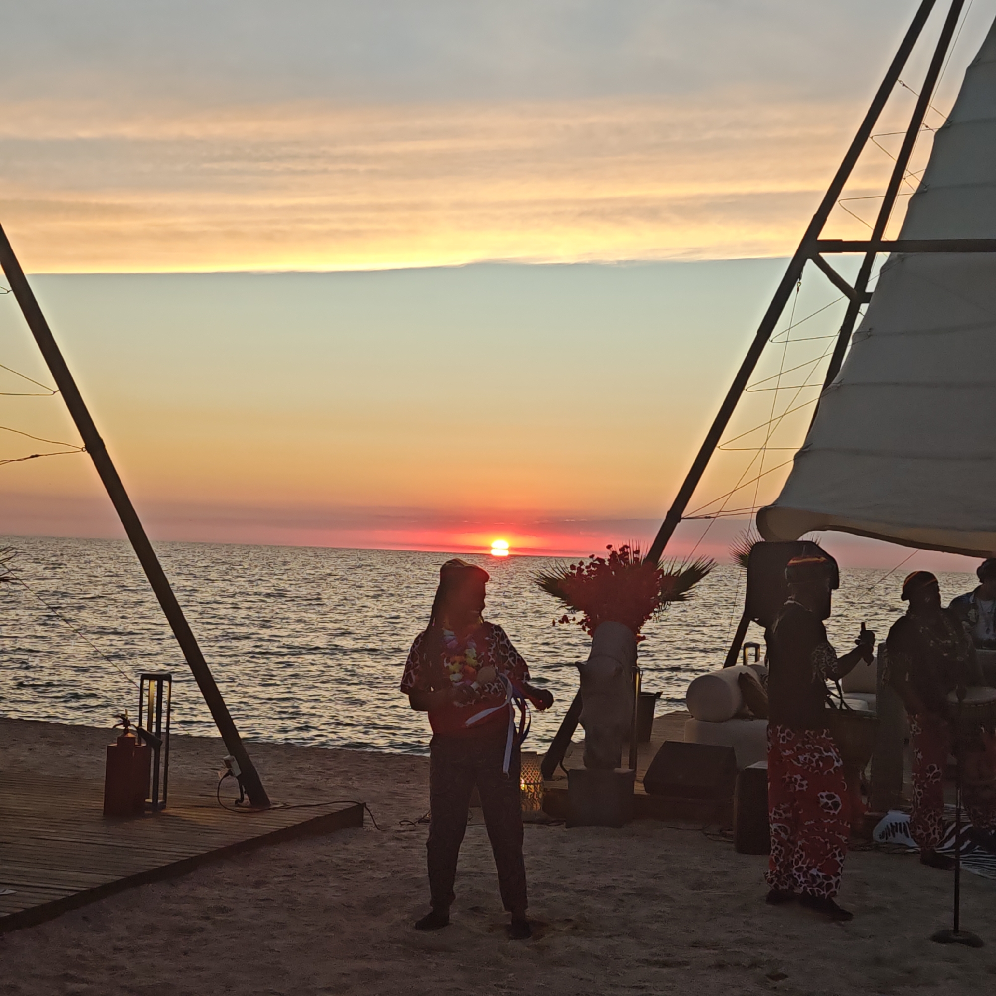 A sunset scene on a beach in Мысовское сельское поселение, Ukraine, features a group of people performing near a body of water. The sun is setting over the ocean, casting a warm glow across the sky and water. Silhouettes of two large mast-like structures, possibly part of a sailboat or temporary stage, frame the scene. In the foreground, on a wooden platform on the sand, several individuals are engaged in what appears to be a musical performance. One person, in the center, wears a colorful lei and a patterned shirt and pants, and is holding a musical instrument, possibly a ukulele. To their right, other individuals are visible, some with drums, suggesting a live band. The environment is sandy, with the ocean providing a backdrop. The lighting is dim, characteristic of twilight.
