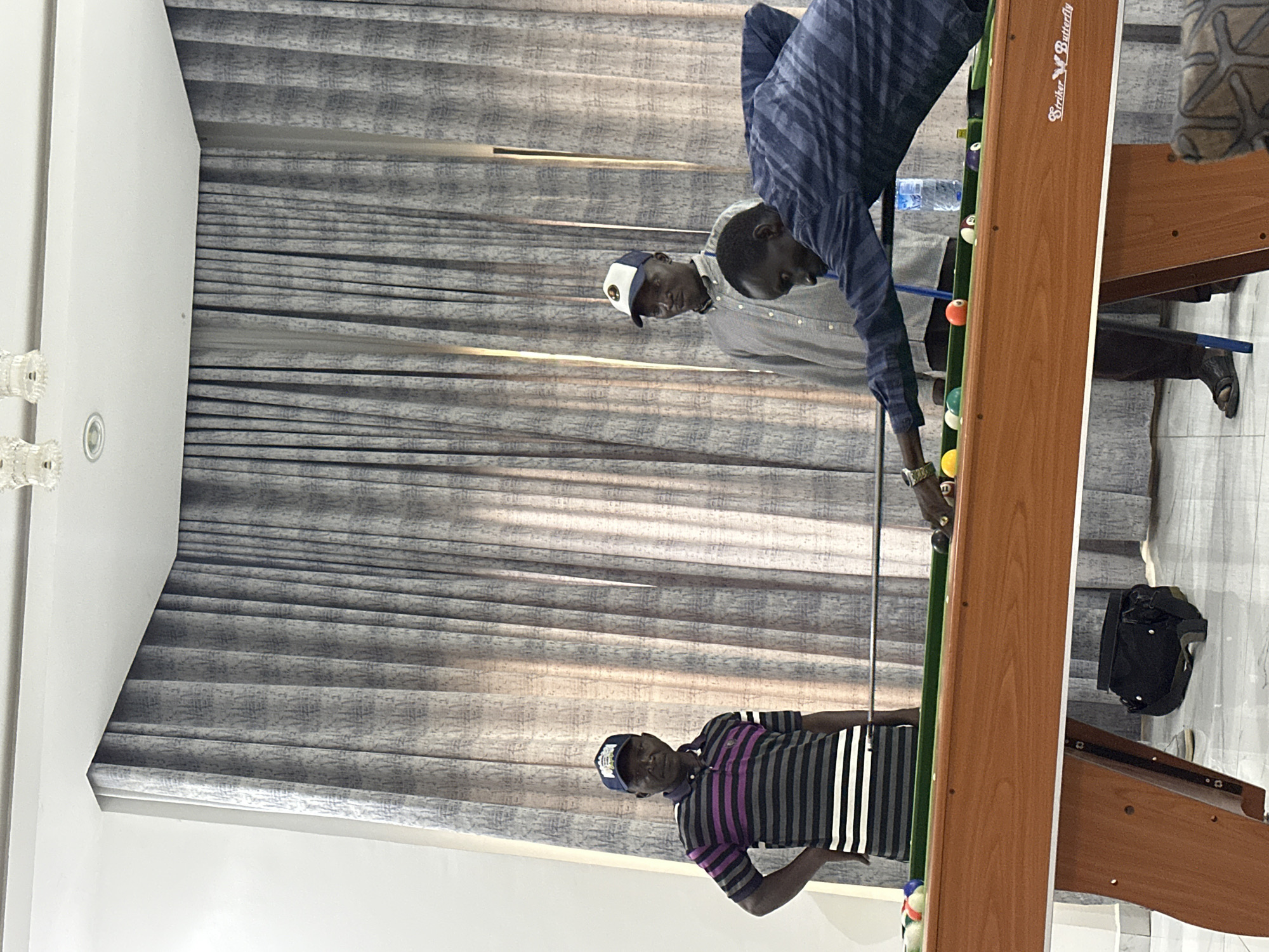 This media file shows three men in a room with patterned grey blinds and white walls. A pool table, labeled "Striker Butterfly," is the central object.
The man in the foreground, wearing a striped shirt and a blue baseball cap, stands near the end of the pool table. He is looking towards the table.
In the middle ground, another man, wearing a checkered shirt and a white and blue baseball cap, is positioned behind the pool table. He appears to be observing the game.
The third man, closest to the pool table, is wearing a blue and white striped long-sleeved shirt. He is actively playing pool, holding a cue stick and aiming at a ball on the table. There are several pool balls visible on the green felt of the table. A bottle of water is also on the table.
The overall lighting suggests it is daytime. The scene appears to be an indoor recreational space, possibly a home or a private club, in Unguwar Sarki, Nigeria. The mood seems relaxed and casual, with the men engaged in a game of pool.