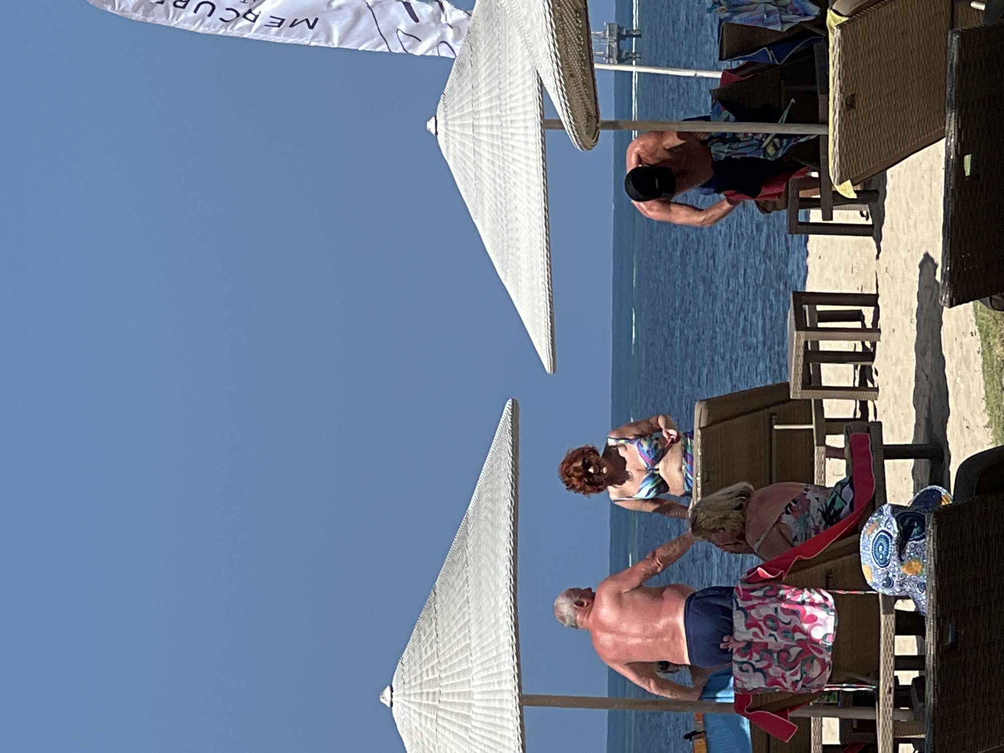 On a sunny day at the beach in Denpasar, Indonesia, several people are enjoying the coastal environment. The scene is dominated by two large, white thatched umbrellas providing shade over beach chairs. The ocean, a clear blue, stretches to the horizon under a bright blue sky.
In the foreground, a man with graying hair and tanned skin, wearing navy blue swim trunks, stands with his back to the viewer. He appears to be interacting with a woman in a colorful bikini who is also facing away from the camera. To the right, another man, wearing a black baseball cap and dark swim trunks, is bent over, appearing to adjust his clothing or belongings.
In front of the beach chairs, a woman with blonde hair, wearing a patterned top, is seated and looking to her left. Another person, wearing a patterned sun hat, is seated in a chair in the lower foreground, facing away from the camera. Colorful towels with floral prints are draped over some of the beach chairs. A white banner with the partial text "MERCURE" is visible in the upper right corner, suggesting the presence of a resort or hotel. The sandy beach is visible beneath the umbrellas and chairs, with some vegetation in the lower left corner. The overall atmosphere is relaxed and recreational, typical of a beach day.