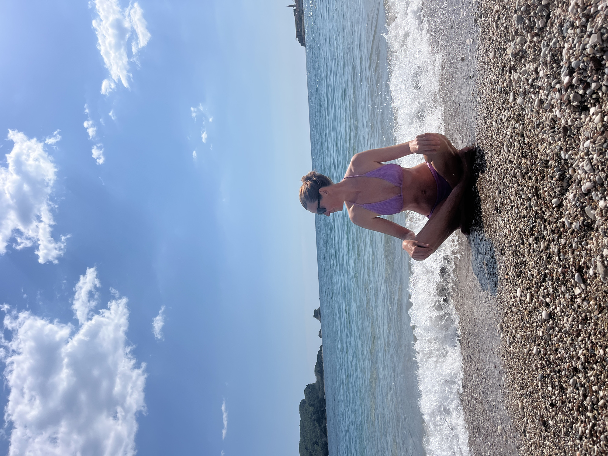 The image captures a woman in a bikini sitting on a pebble beach in Budva, Montenegro. The woman is seated cross-legged at the edge of the water, with waves washing around her. The beach is composed of small, rounded pebbles. The sea stretches out behind her, and in the distance, a rocky headland can be seen. The sky is blue with scattered white clouds, indicating a sunny day.