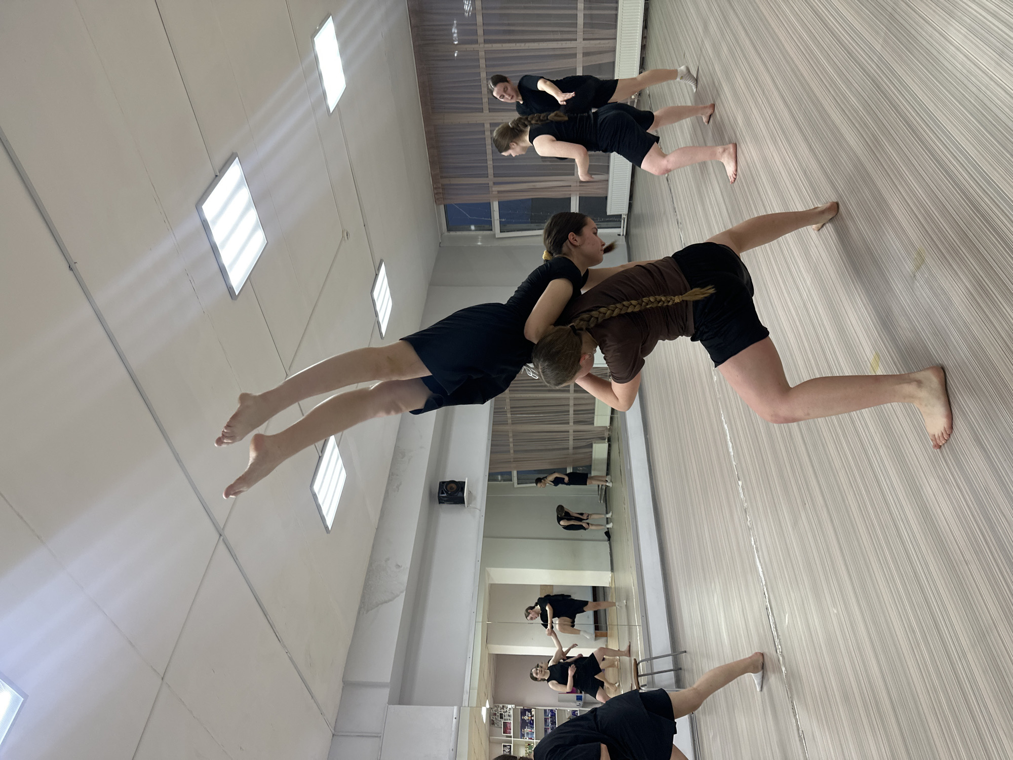 The image shows a dance studio in Votkinsk, Russia. Several young women in dance attire are practicing. Two women in the foreground are performing a partner acrobatic pose, one supporting the other on her shoulders. Other dancers are practicing in the background and in the mirror reflection. The studio has a light-colored floor and ceiling with rectangular light fixtures. A large mirror covers one wall.