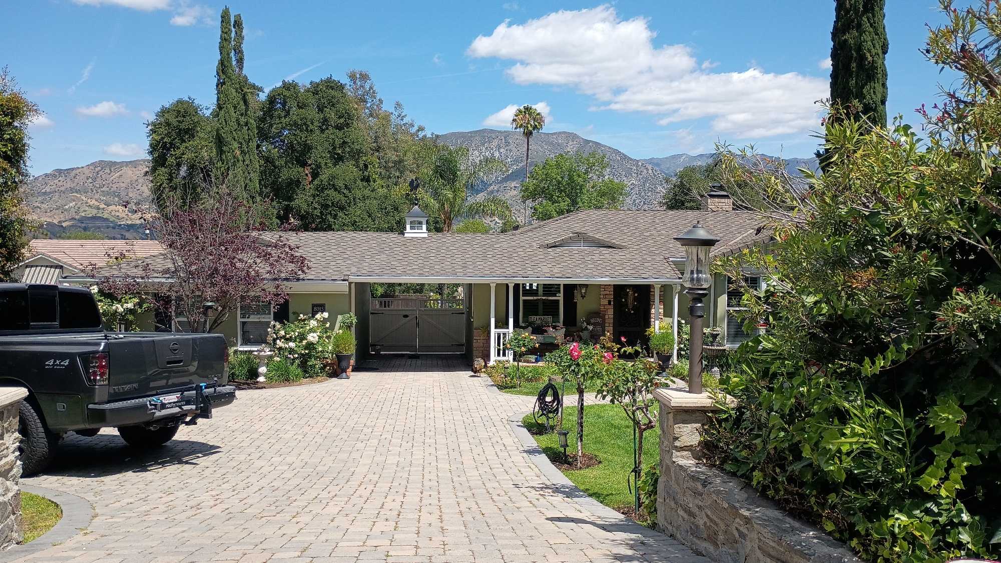 This media file captures a sunny daytime view of a well-maintained single-story house with a paved driveway, set against a backdrop of mountains under a clear blue sky with scattered white clouds. The scene conveys a peaceful, residential atmosphere, likely in the Los Angeles area.
The house features a light green or tan exterior and a brown shingled roof. A small white cupola with a weather vane is visible on the roofline. A covered porch with pillars is adorned with potted plants and rose bushes with pink blossoms. A sign on the porch, partially obscured, reads "FLEA MARKET." The house number "8015" is visible near the front left corner. To the left, large, light-colored gates open into a covered carport or drive-through area.
The curved driveway is constructed from light-colored interlocking pavers. Partially visible on the left side of the driveway is the rear of a dark grey Nissan Titan pickup truck. The truck's branding "4x4" and "TITAN" are clearly legible. A bike rack, branded "rockymounts," is attached to the truck's rear hitch.
The landscaping is rich and varied, featuring well-manicured lawns, several rose bushes with both pink and white flowers, and numerous other plants and shrubs. Tall, slender evergreen trees stand prominently, while a distant palm tree adds to the natural scenery. A low stone wall with a matching stone pillar and an outdoor lamp post marks the edge of the property in the foreground. The background is dominated by rugged mountains, typical of the Los Angeles landscape, covered in natural vegetation.