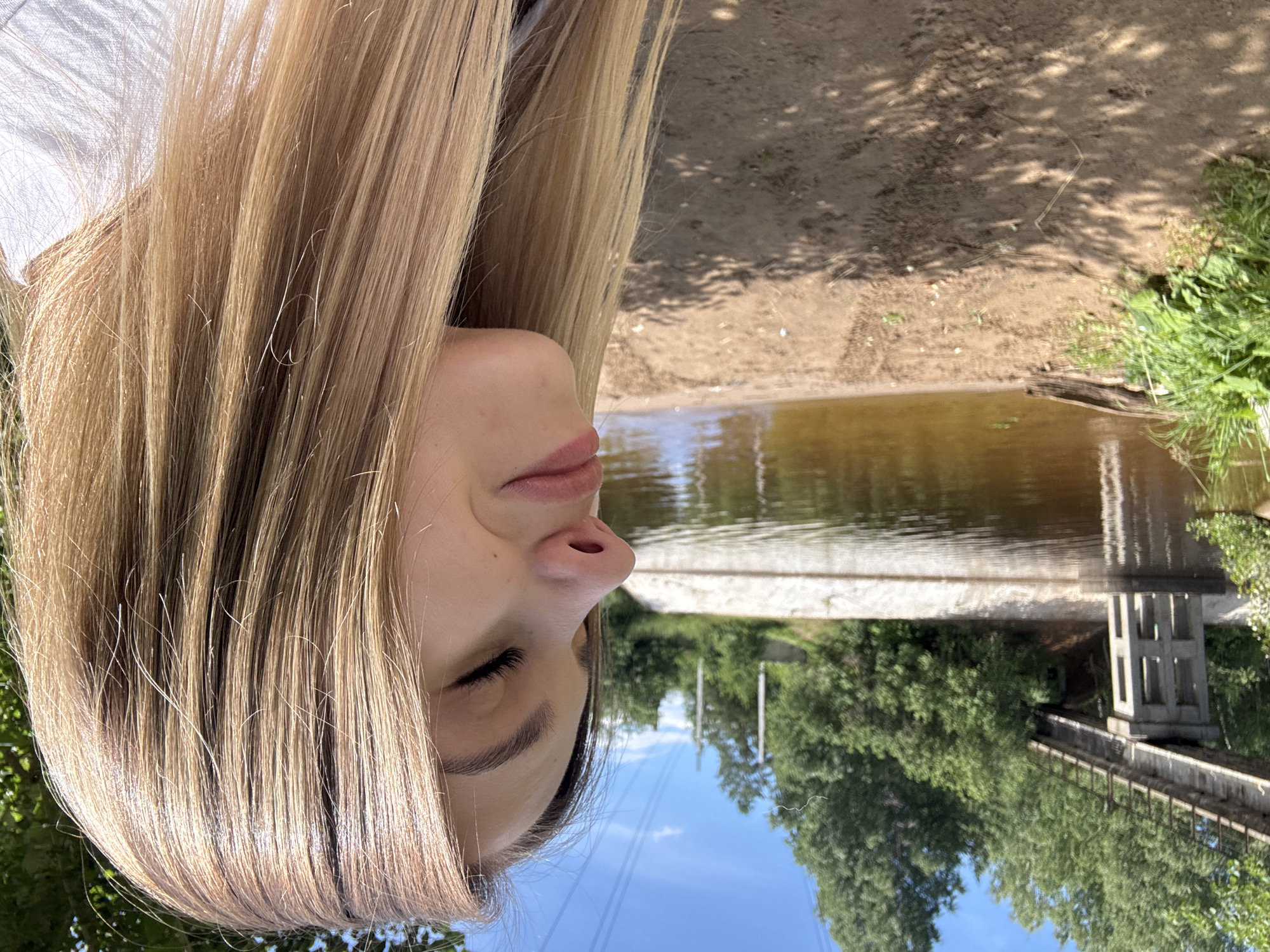 This is an upside-down, close-up selfie of a young woman with long, blonde hair. Only her face, from the nose down to her chin, and her hair are visible. She is looking down and to her right, with her eyes closed and a slight smile on her lips. Her hair is neatly parted and cascades over her face and shoulders.
The background suggests an outdoor setting, likely near a body of water. The water appears calm and reflects the surroundings. Behind the water, there are trees and a concrete structure that resembles a bridge or a dam. The ground is sandy with some scattered vegetation. The lighting indicates it is daytime, with bright sunlight casting shadows. The overall mood is relaxed and peaceful, possibly indicating a moment of personal reflection or enjoyment of nature. The location context is Chaikovsky, Russia. There is no visible text in the image.