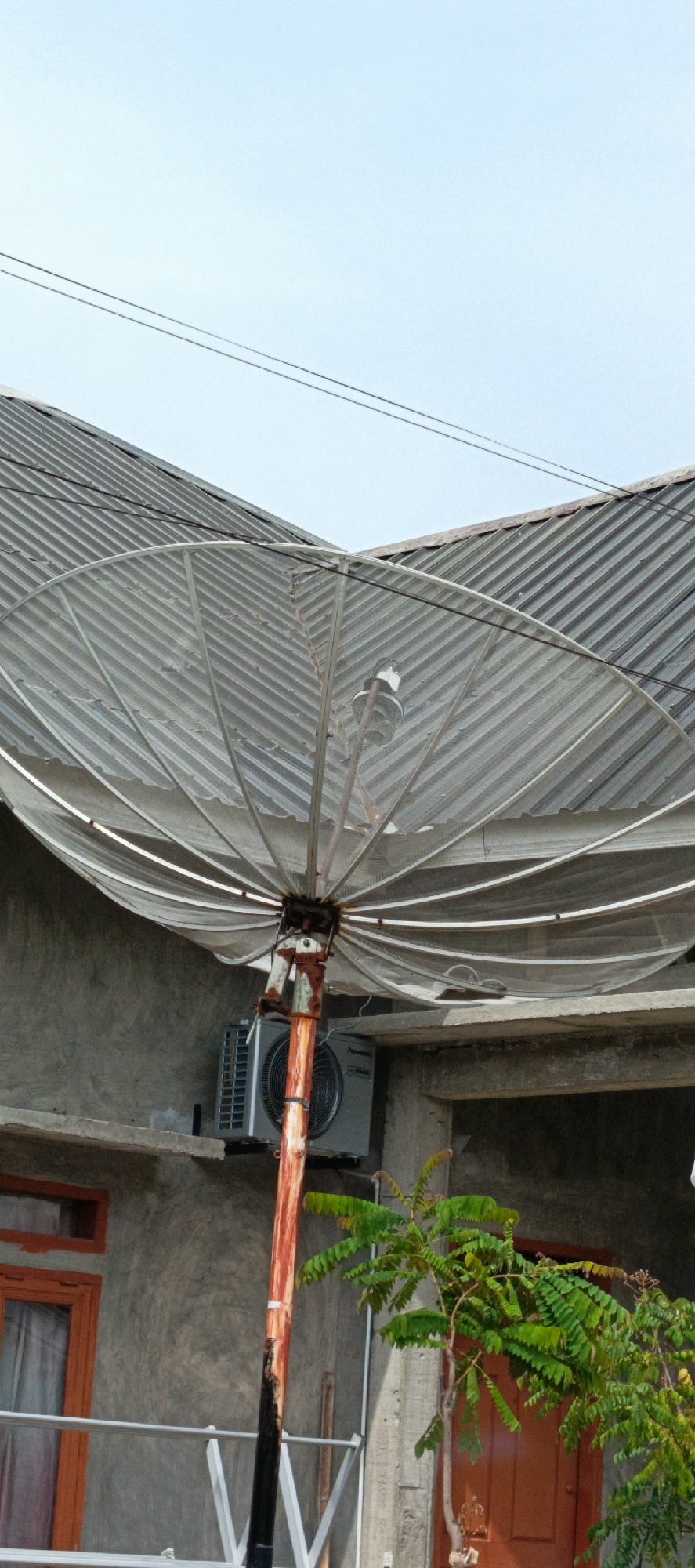 This image depicts an outdoor scene, likely in a residential area of Pasi Rawa, Indonesia, focusing on a satellite dish and surrounding structures. A large, weathered satellite dish, made of a mesh-like material, is prominently displayed. It is mounted on a rusty, reddish-brown pole that shows signs of wear and tear. Behind the satellite dish, a corrugated metal roof of a building is visible, angled against a clear blue sky.
To the left of the satellite dish, an air conditioning unit is mounted on a concrete ledge, attached to a grey-textured wall. A window with an orange frame is also visible on this wall. In the lower right portion of the image, a small tree with vibrant green leaves is growing, adding a touch of nature to the scene. A brown door is partially visible behind the tree. The overall impression is that of a functional but somewhat aged outdoor space in an Indonesian locality.