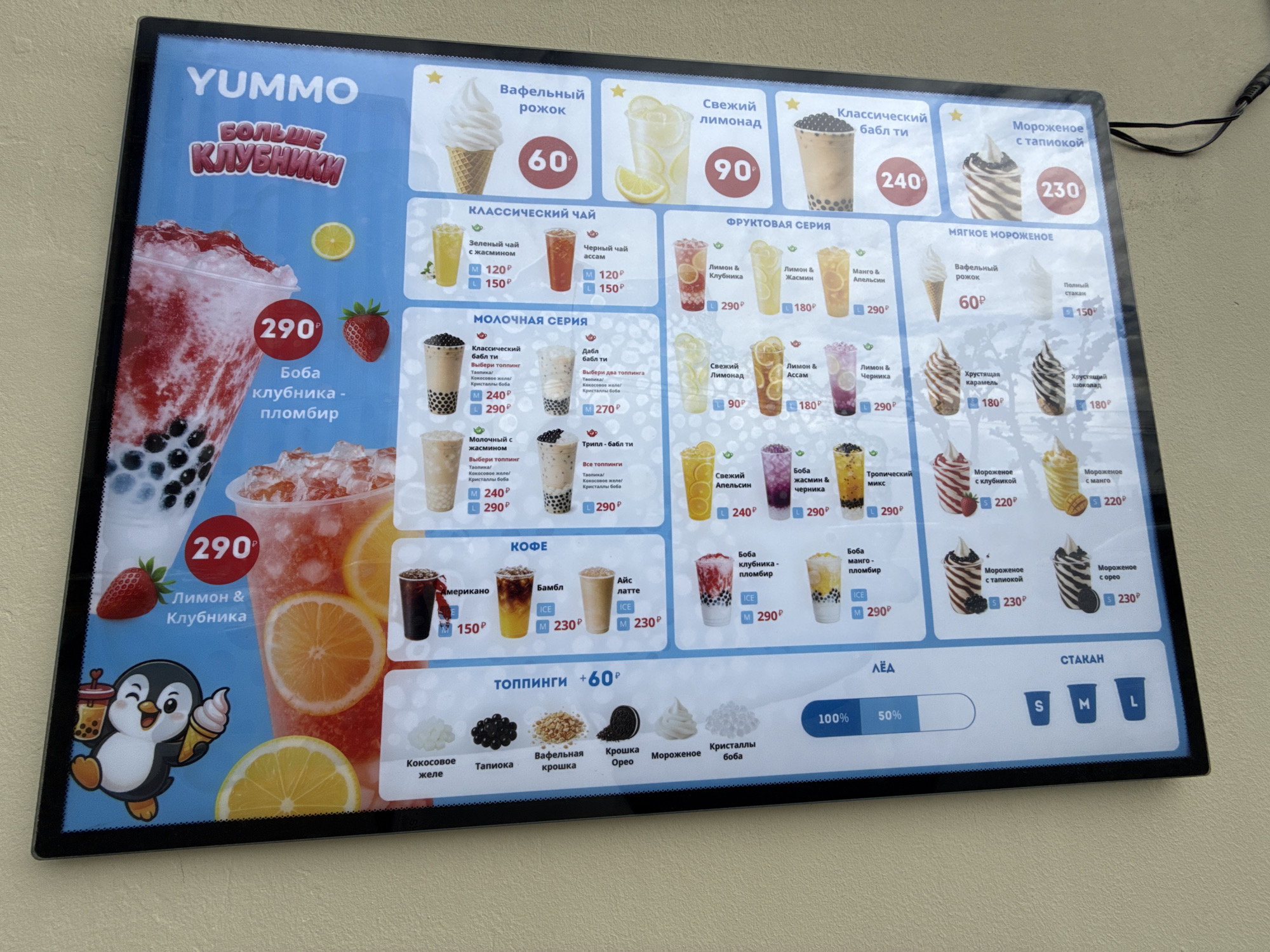 This is a close-up, eye-level shot of a brightly lit digital menu board for a dessert shop called "YUMMO". The menu features a variety of ice cream, bubble tea, and coffee drinks. The dominant colors are a vibrant sky blue and pastel shades of pink, yellow, and white, creating a cheerful and inviting atmosphere.
The menu is divided into sections, with prominent images of the featured items. A large illustration of a strawberry and lemon drink with the price "290" is positioned on the left, accompanied by a cartoon penguin holding an ice cream cone and a bubble tea. This suggests a playful and fun brand identity.
Several sections showcase different drink categories: "Classical Tea", "Fruit Series", "Milk Series", and "Coffee". Within these, there are pictures of various bubble teas, iced coffees, and other beverages, each with their prices in rubles, denoted by "P". The prices range from 60P for a waffle cone to 290P for some of the more elaborate bubble teas.
A dedicated section for "Toppings" (+60P) displays small images of ingredients like coconut jelly, tapioca, crushed Oreos, and cream. Below this, there are options for "Ice" and "Glass" sizes (S, M, L).
The overall impression is a modern, visually appealing menu board designed to attract customers with its colorful presentation and clear pricing. The lack of people in the frame focuses all attention on the tempting food and drink offerings. The lighting suggests an indoor setting, likely a shop or cafe, possibly during the daytime given the bright illumination. There are no obvious weather cues or specific Moscow landmarks visible, making the location context primarily inferred from the Cyrillic text and common business practices.