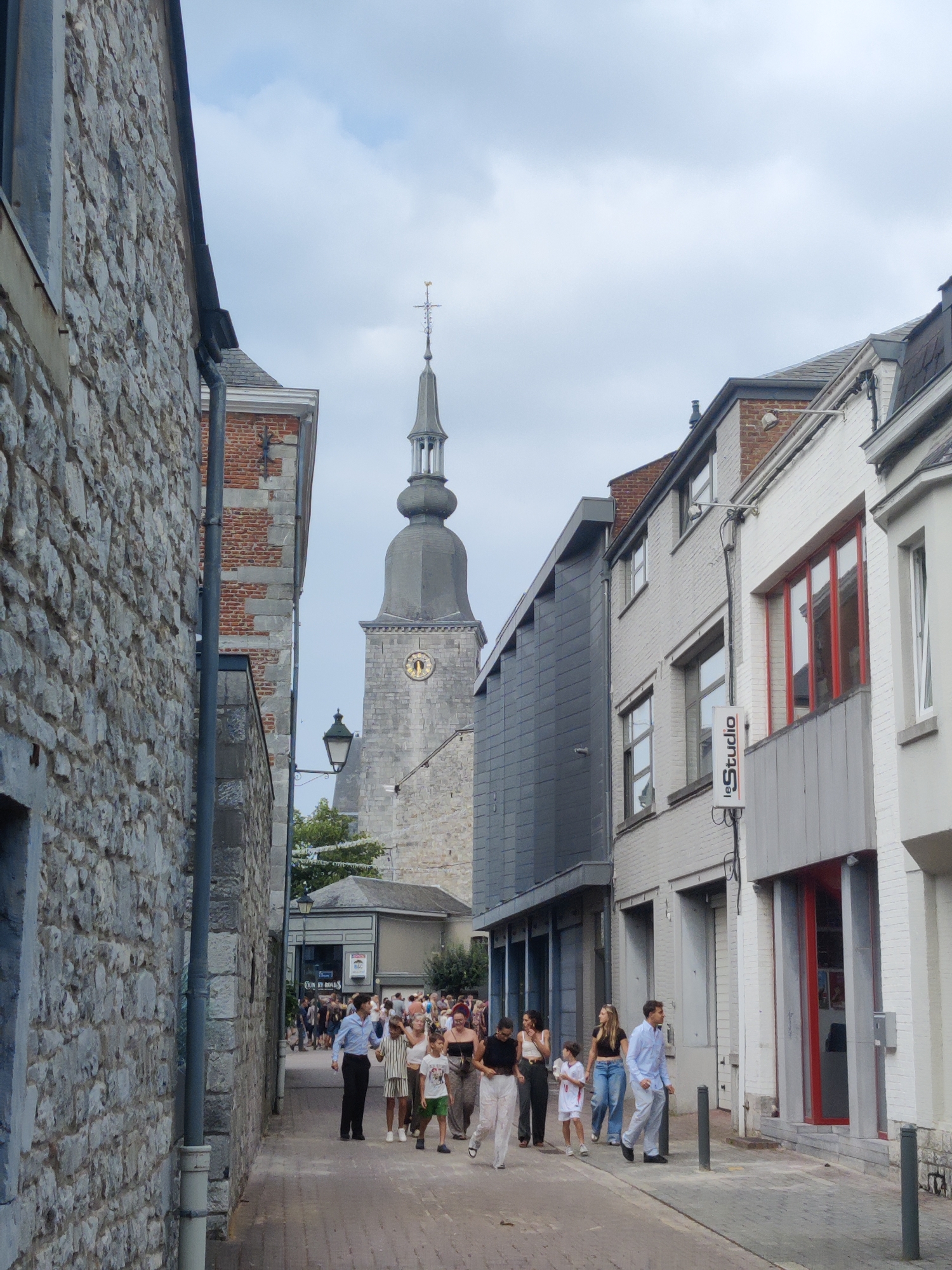 In Marche-en-Famenne, Belgium, a street scene unfolds with a stone-walled building on the left and a row of lighter-colored buildings on the right. In the distance, a tall clock tower with a spire dominates the skyline. A group of people are walking down the cobblestone street, some in casual attire, others dressed more formally. A man in a blue shirt and light-colored pants is walking with a woman and a child. The sky is overcast, with hints of blue peeking through the clouds. The overall atmosphere is one of a pleasant day in a historic European town.