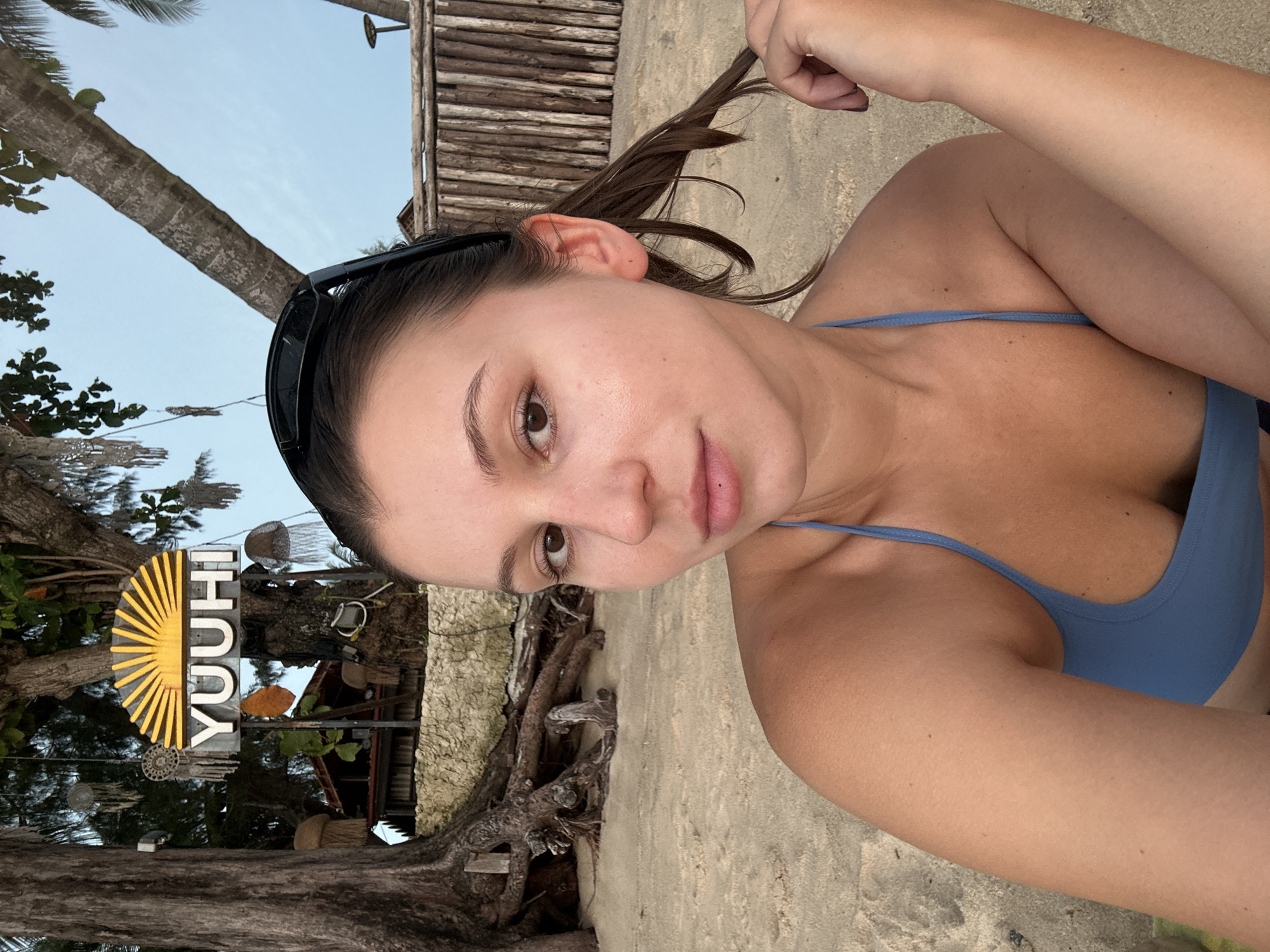 A young woman with brown hair pulled back and sunglasses perched on her head is seated on a sandy beach, looking towards the camera. She is wearing a blue bikini top. Behind her, a sign with the word "YUUHI" and a sunburst design is visible, attached to a tree with exposed roots. The background includes palm trees, a wooden fence, and a clear sky, suggesting a tropical location like Choeng Thale, Thailand.