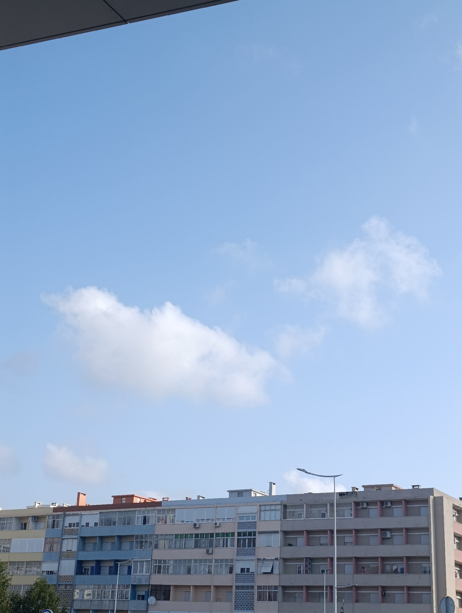 The image shows a view of apartment buildings under a clear blue sky with a few scattered clouds. The buildings are multi-story, with a mix of colored facades, including yellow, blue, and light gray. Many windows and balconies are visible, some with closed shutters or blinds. Air conditioning units are mounted on some of the building exteriors. A modern street lamp with a curved arm stands in front of one of the buildings. The top edge of a dark, angular structure, possibly a building overhang or roofline, is visible in the upper portion of the image. The scene appears to be a residential area in Setúbal, Portugal.