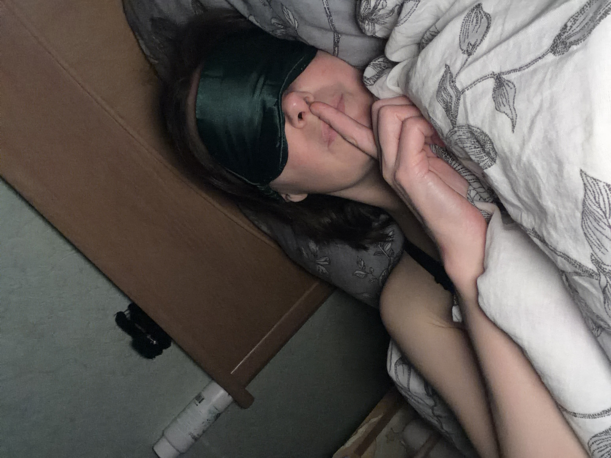 A person wearing a dark green sleep mask is lying in bed, picking their nose. The bed is covered in patterned sheets and a white duvet with a floral pattern. A bedside table is visible to the left, next to a gray wall. A black object and a white tube are on the floor. The location is Unknown, Unknown.