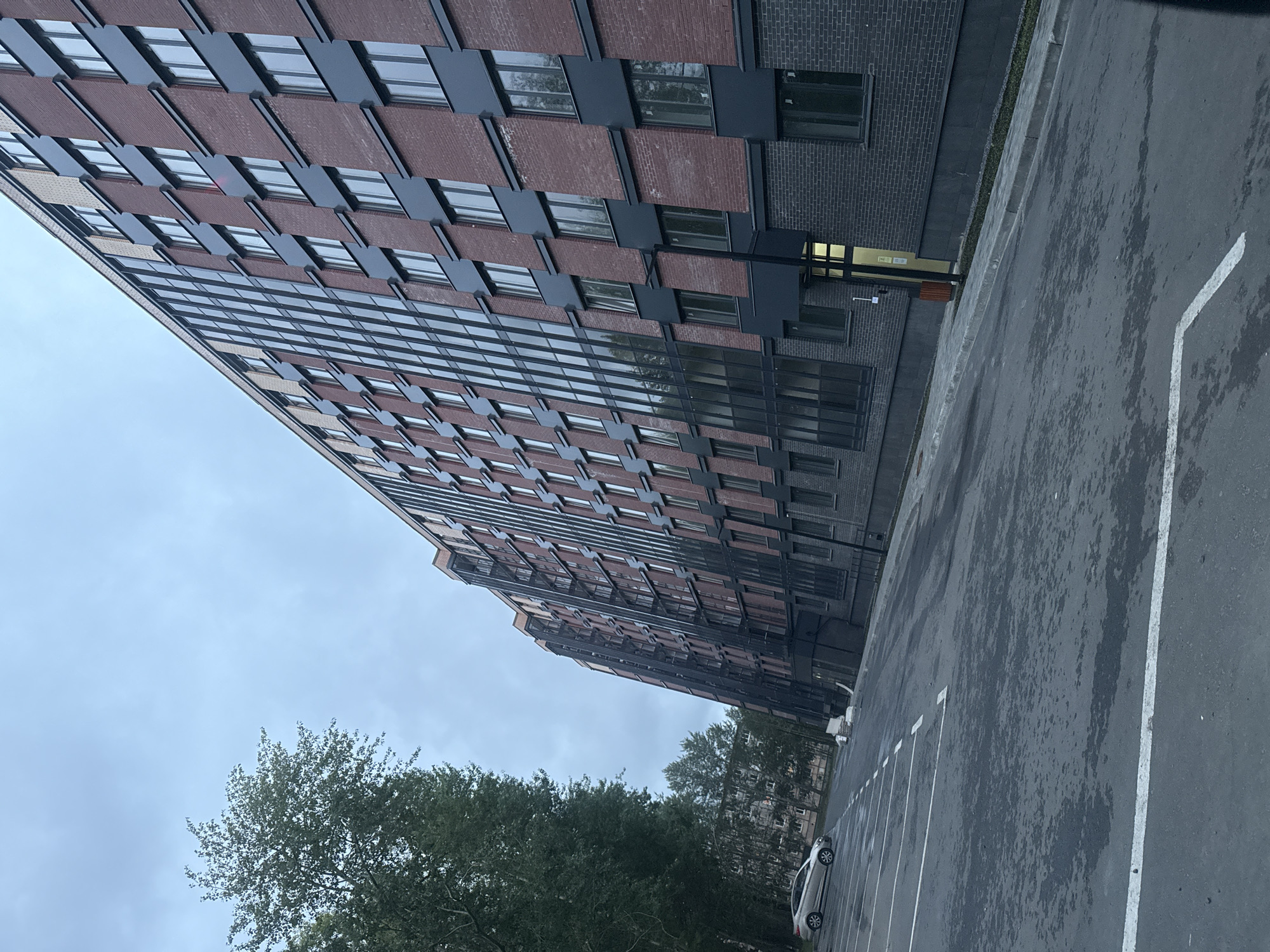 A long, modern, multi-story apartment building with dark brick and glass facade is situated in Severodvinsk, Russia. A parking lot with white lines is in the foreground, with one silver car parked on the left. Trees are to the left of the building, and a cloudy sky is overhead. The building appears to be newly constructed.