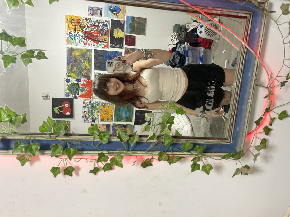 A young woman with brown hair and bangs poses for a mirror selfie. She wears a white tank top and a black skirt, adorned with various rings and bracelets. The ornate mirror frame is decorated with artificial ivy and a red LED light strip. Behind her, the wall displays numerous colorful artworks. A white fan and clothes are seen in the background. The overall scene has a casual, artistic vibe.
