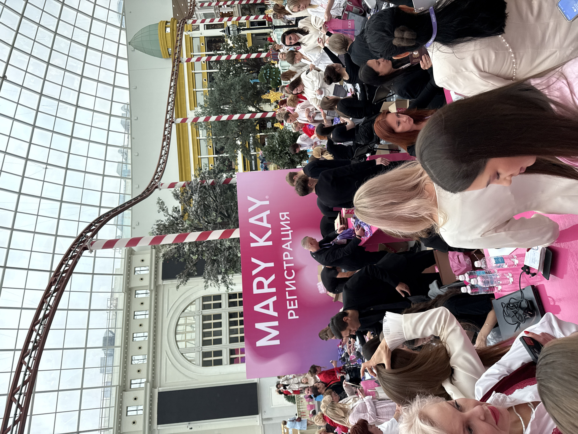 The image depicts an indoor event, likely a promotional or sales gathering, with a large pink banner prominently displaying "MARY KAY" and Russian text that translates to "REGISTRATION." A crowd of people, predominantly women dressed in formal or semi-formal attire, are gathered around tables covered in pink cloths. They appear to be interacting with each other, possibly engaging in registration, receiving products, or attending to presentations.
The environment suggests a large indoor space with a high, glass-paneled ceiling, characteristic of an atrium or a shopping mall. Architectural elements visible in the background include a white building with arched windows and a red, rollercoaster-like structure overhead. Red and white striped poles are also present, possibly for decoration or structural support. Some trees and greenery are visible, indicating an attempt to bring an outdoor feel indoors. The presence of water bottles and what appear to be cosmetic products on the tables suggests a beauty-related event.
Based on the visual cues, this scene is likely taking place in an event space within a large shopping mall or exhibition center in Moscow, Russia. The architecture and scale are consistent with modern large-scale retail and entertainment complexes found in the city.