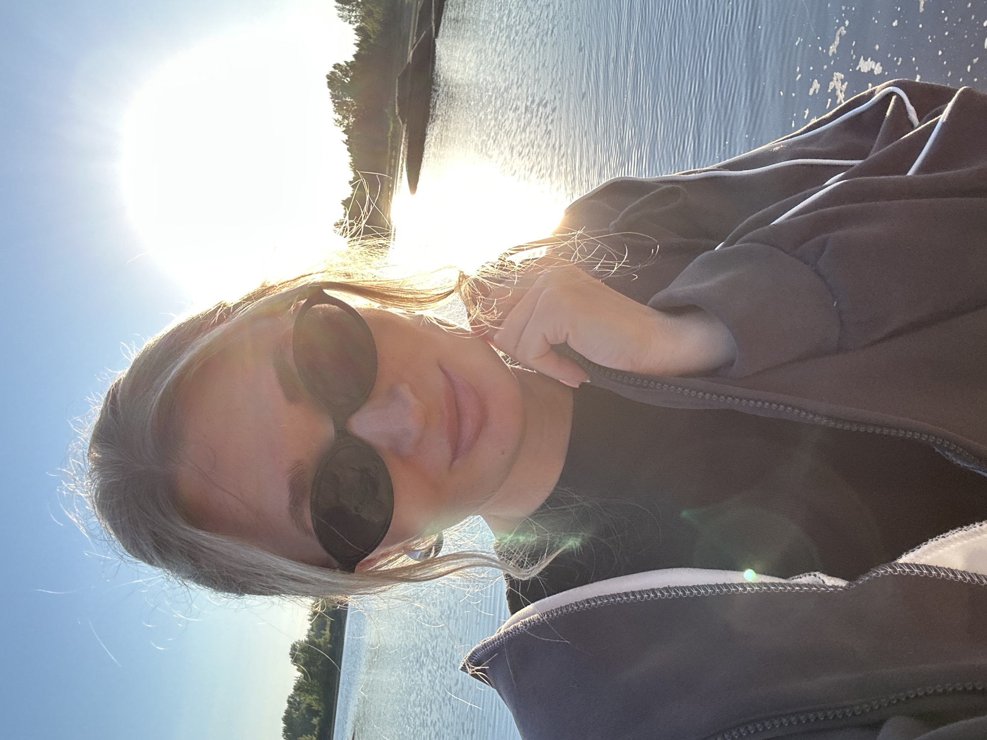 A young woman with blonde hair is taking a selfie outdoors by a body of water, possibly a river or lake, with the sun setting behind her, creating a bright glare. She is wearing sunglasses and a dark jacket over a black shirt. The background shows a tree line and the surface of the water. The lighting suggests it is either sunrise or sunset. The image has a warm, natural light.