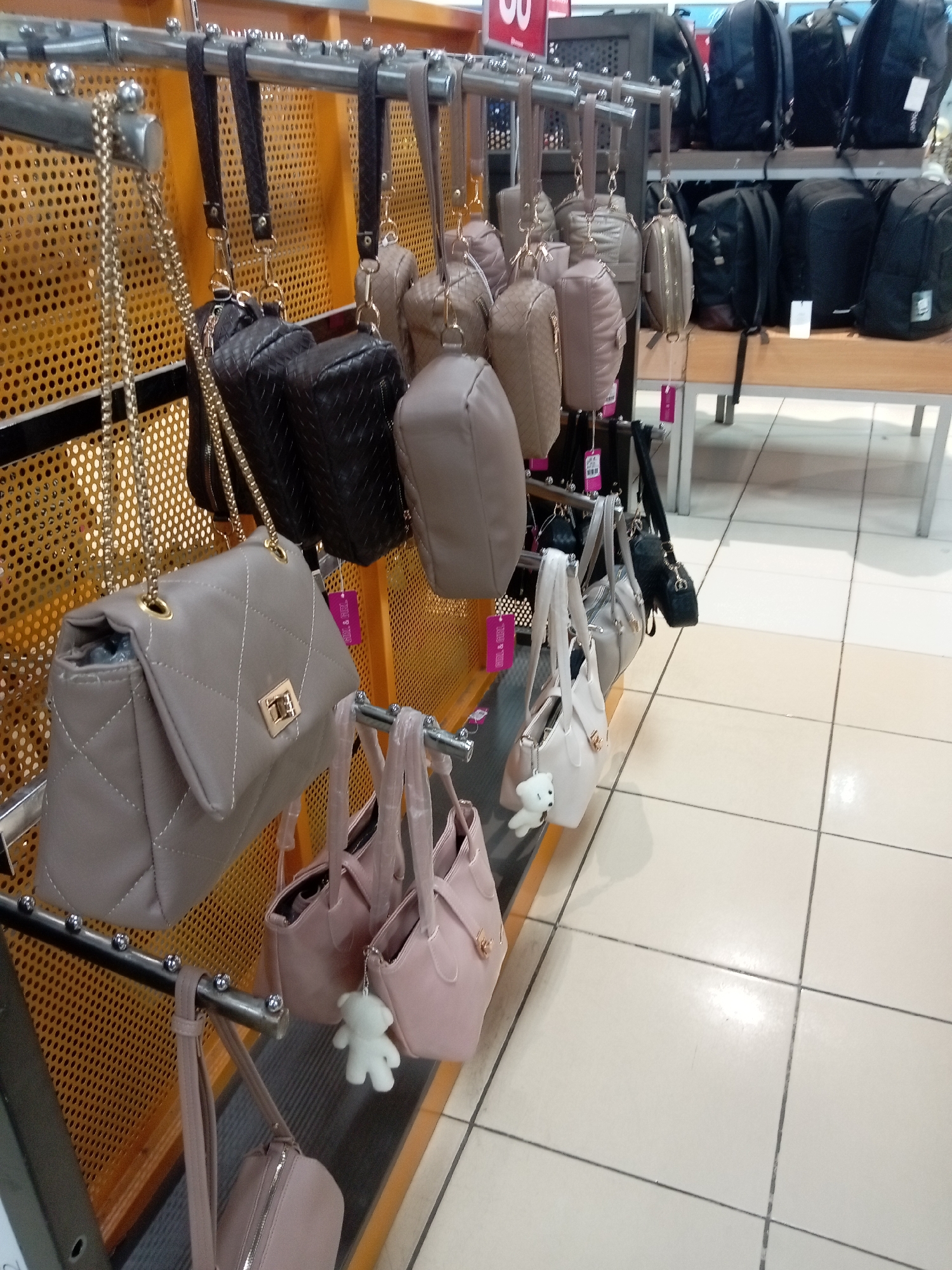 The image displays a retail store setting, likely a handbag or accessory shop, showcasing a variety of handbags displayed on metal racks. The store appears to be located in Batam, Indonesia, suggested by the overall aesthetic commonly found in Indonesian retail environments and the specific products on display which cater to local fashion trends.
In the foreground, several handbags are prominently featured, hanging from metallic rods. The handbags vary in style, color, and material. One prominent handbag, in a light taupe color, is quilted with white stitching and features a gold clasp. It hangs from a long, delicate gold chain. Adjacent to it, and slightly behind, are two dark brown, woven-patterned handbags. Further down the rack, there are several smaller, rounded handbags in various shades of beige and taupe, some adorned with teddy bear charms. At the bottom of the rack, a pink handbag with a long strap is visible.
In the background, more handbags are visible on higher racks, including a collection of black and dark grey backpacks. The walls behind the display racks are a textured, perforated orange material, typical of retail fixtures designed for hanging merchandise. The floor is tiled in a light cream color with visible grout lines. The lighting appears to be bright, typical of an indoor retail environment. No people are visible in the image. The overall impression is one of organized product display in a commercial space.