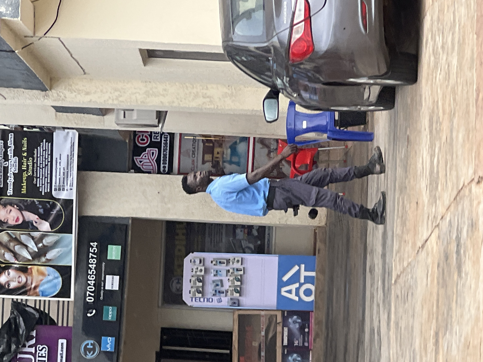 A Black man wearing a light blue short-sleeved button-down shirt and dark grey pants walks past a row of shops in Okpanam, Nigeria. The man is wearing black boots and is looking to his left. Behind him, a dark grey car is parked. The shops have various signs and displays, including a makeup and nails studio and a display of mobile phones. The pavement is concrete.