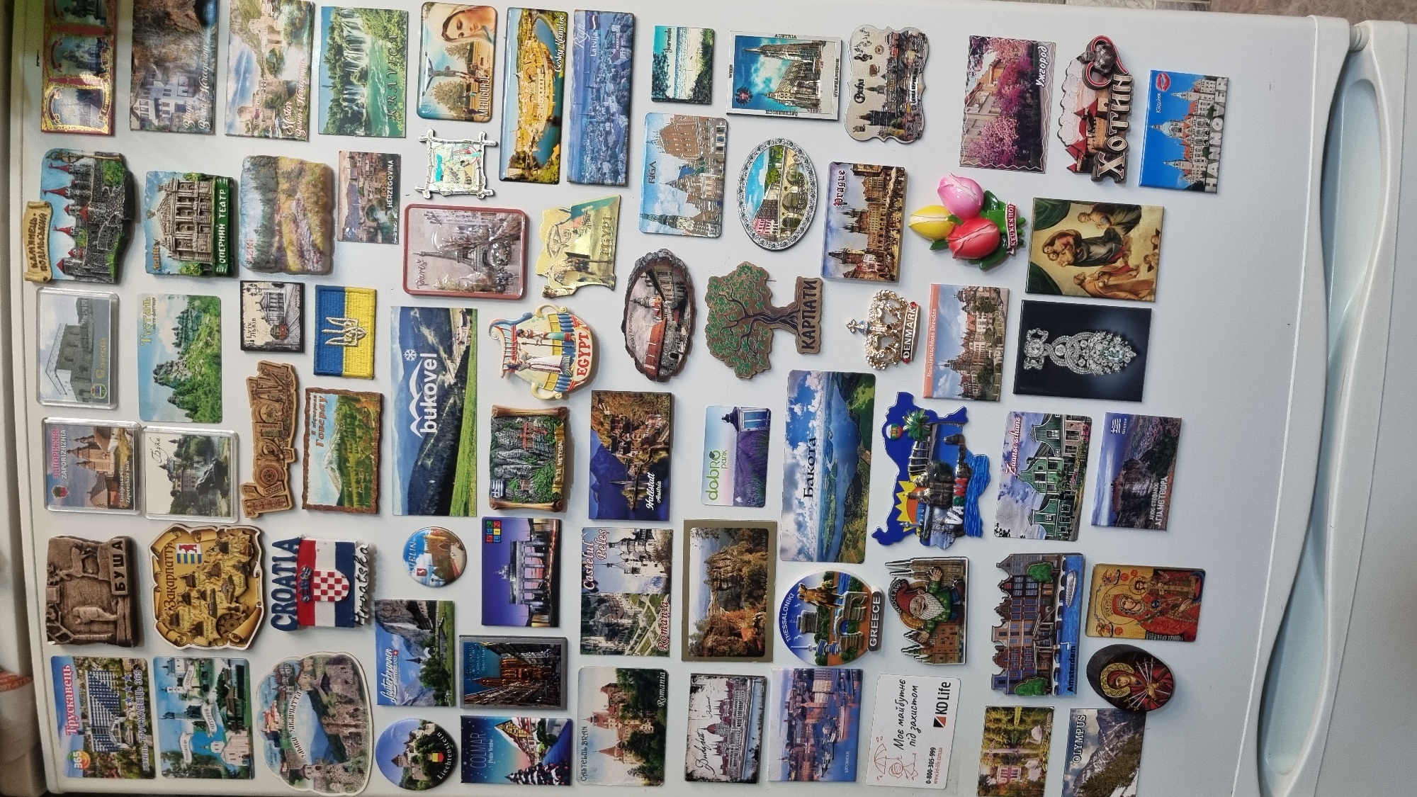 The image displays a white refrigerator door completely covered with a variety of magnets, predominantly depicting landmarks, maps, and scenic views from different countries and cities. Many of the magnets are rectangular or irregularly shaped, featuring colorful illustrations and text. Some of the identifiable locations include Croatia, Egypt, Greece, Austria, Germany (Berlin and Dresden), Latvia (Riga), and various Ukrainian locations such as Bukovel and Karpati. There are also magnets with images of religious art, architectural structures, and natural landscapes. The overall impression is that of a collection of travel souvenirs. There is no specific visual information present in the image that allows for the identification of the city of Smila, Ukraine, or any specific details related to that location. The scene is static and shows no people or actions.
