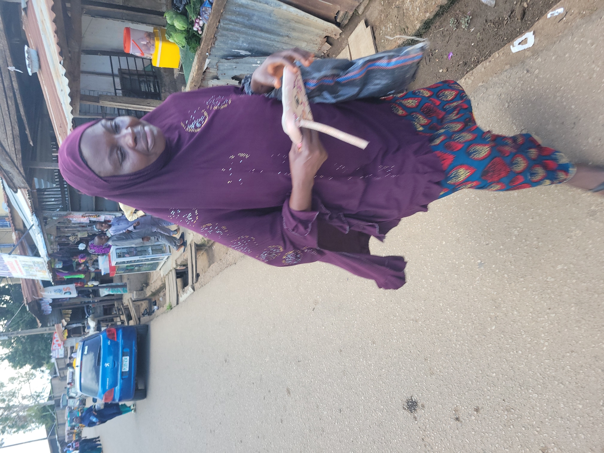 A woman wearing a purple hijab and a patterned skirt walks on a paved road in what appears to be a marketplace in Jos, Nigeria. The woman is holding a piece of paper or thin object in her right hand and a dark plastic bag in her left. To her right, there are market stalls with various items, including vegetables and containers. A blue Peugeot car is parked on the side of the road behind her. In the background, other people and market structures are visible, indicating a busy urban environment. The overall scene depicts a typical day in a bustling Nigerian city.