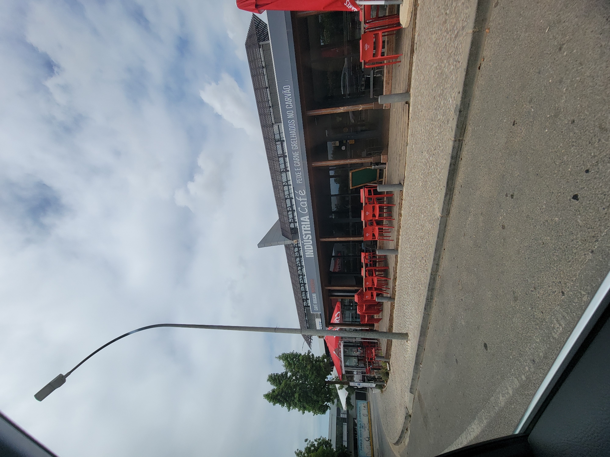The image depicts a street-level view of "INDUSTRIA CAFÉ PEIXE E CARNE GRELHADOS NO CARVÃO" (Industry Café Grilled Fish and Meat on Charcoal), located in Loulé, Portugal. The establishment features an outdoor seating area with numerous red chairs arranged around tables, some covered by a red umbrella. The café itself has large glass windows and a wooden deck in front. A street lamp stands tall to the left of the building. In the background, trees and other buildings are visible. The scene appears to be a daytime shot under a cloudy sky.