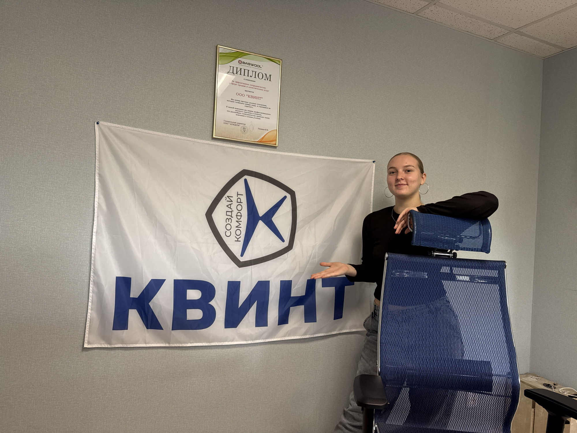 A young woman with blonde hair tied back and wearing a black long-sleeved shirt and jeans stands in front of a white flag with a blue logo and text on it. The flag has the word "КВИНТ" in large blue letters. The logo on the flag is a black hexagonal shape with a blue star-like symbol inside and the words "СОЗДАЙ КОМФОРТ" written in Russian. To the left of the woman, on the wall, hangs a framed certificate with Russian text. The woman is leaning on a blue mesh office chair. The environment appears to be an office with grey textured walls and a dropped ceiling with fluorescent lighting. A cardboard box is visible on the floor to the right. The overall scene depicts an employee posing with company branding in what is likely an office space.