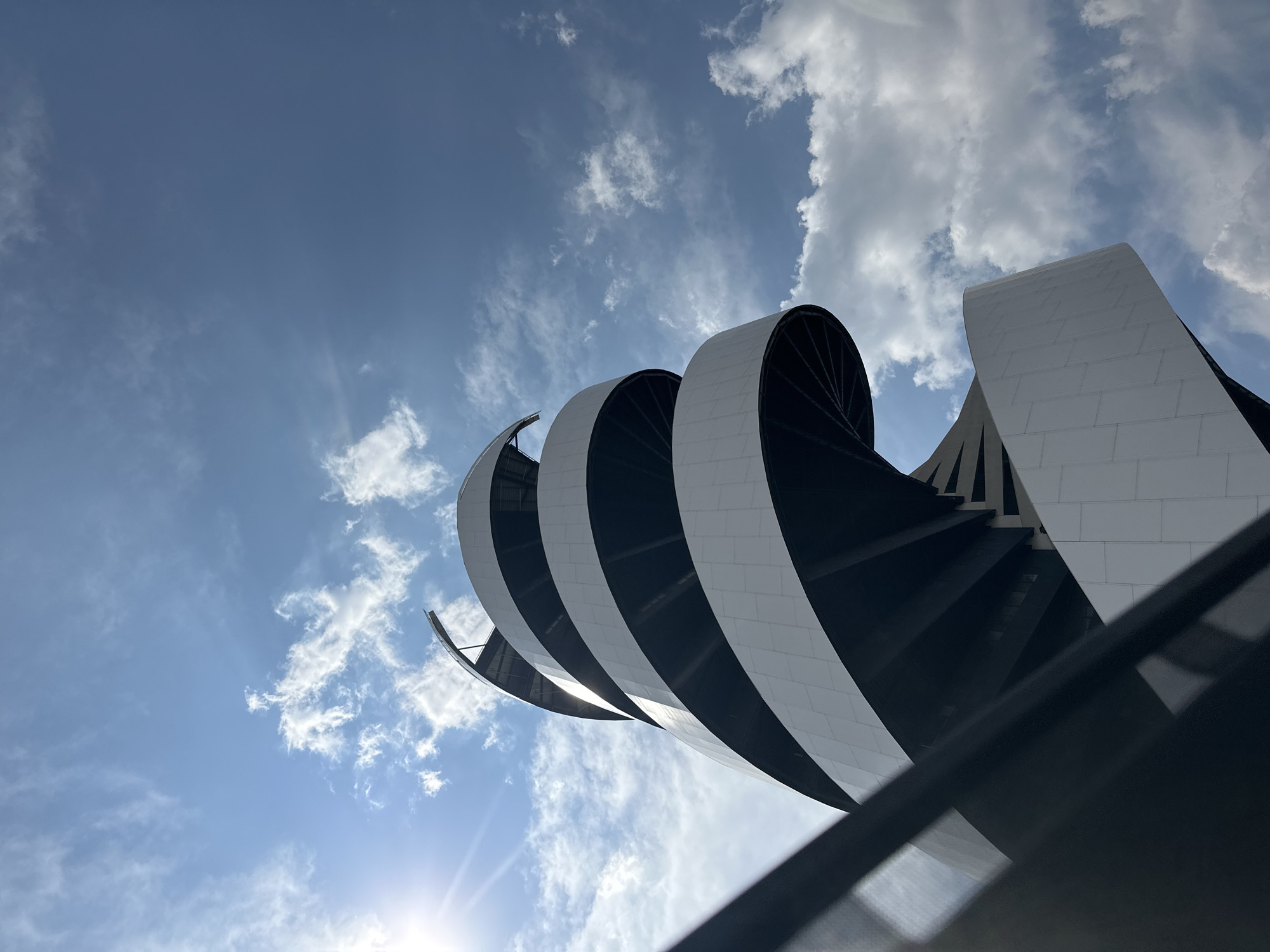 This is a low-angle architectural photograph showing a section of a building with a distinctive, repeating curved or spiral design. The structure consists of a series of white and dark-colored segments that arch and appear to stack upon each other, receding into the frame. The sky fills the background, featuring a mix of blue sky with sunlight and scattered clouds. A low structure, presumably a barrier or handrail is partially visible at the bottom edge of the image. The location is in San Bernardino Tlaxcalancingo, Mexico.