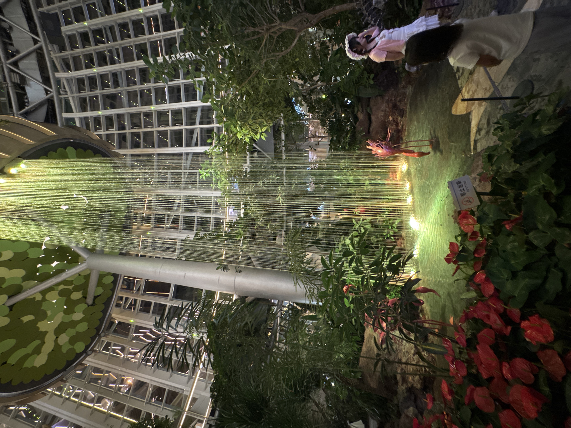 The image shows an indoor botanical garden with a waterfall of light strands cascading down from a tree-like structure. The surrounding area is filled with lush green plants and red flowers. A decorative flamingo statue stands in the water. Two people, a woman in a white dress and a pink cardigan, and a child in a white t-shirt, are visible in the scene. The location is identified as Liangjiang, China. The environment appears to be a large, modern building with a glass-paneled ceiling and walls, allowing natural light to filter in, though the image is taken at night or in low light conditions, with artificial lighting illuminating the waterfall and plants. There are also signs in the foreground, one of which reads "NO SITTING" in English.