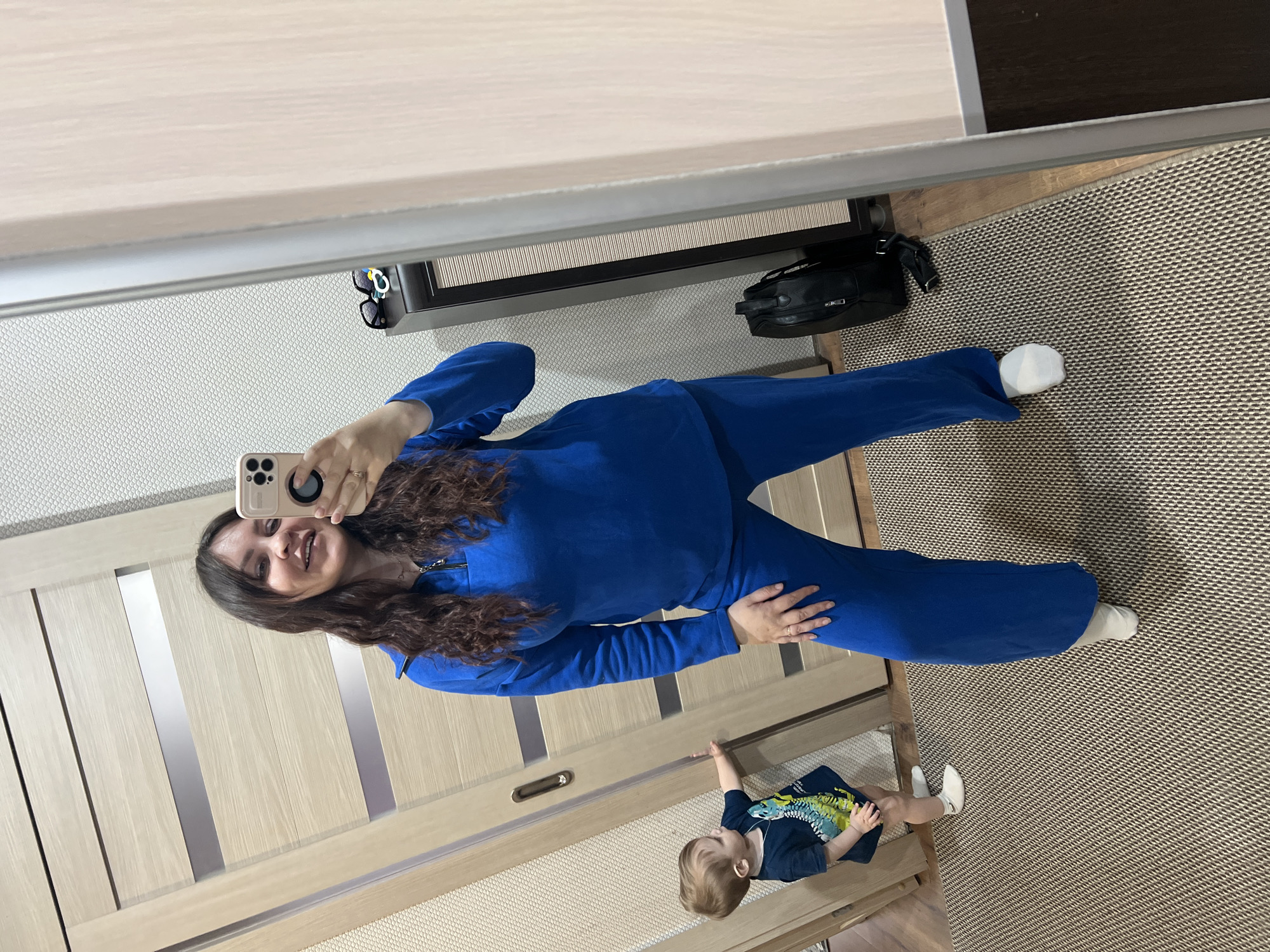 A woman in a blue tracksuit is taking a selfie in a mirror. A young child in a navy blue shirt with a green and yellow design is near her feet. The background includes a light wood-paneled door, a patterned rug, and a closet with items like sunglasses and a black bag on the top shelf. The location, Noviy Urengoy, Russia, is assumed to be implied.