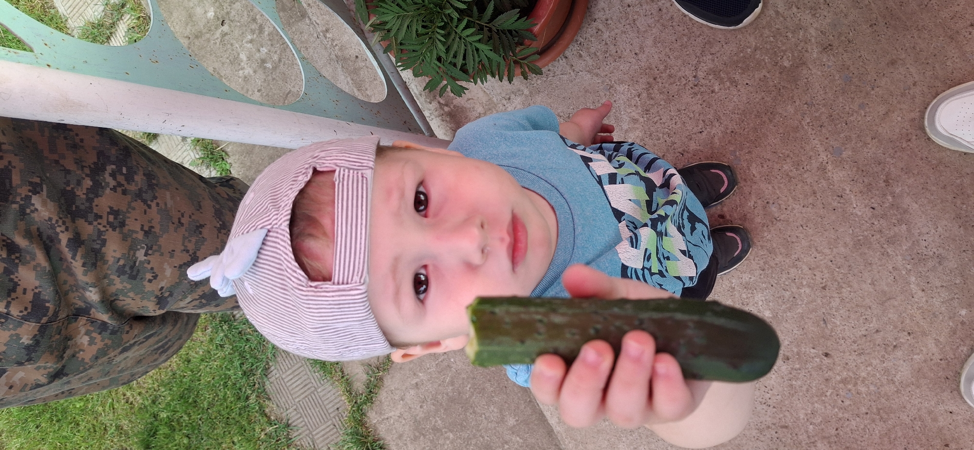 A child, wearing a striped cap with small bunny ears, looks up at the camera while holding a piece of cucumber. The child has rosy cheeks and is wearing a blue shirt with a colorful pattern. The background shows a concrete ground and some green grass, suggesting an outdoor setting. The image appears to be taken during the day. There are also glimpses of other people's shoes in the frame, indicating they are present with the child.