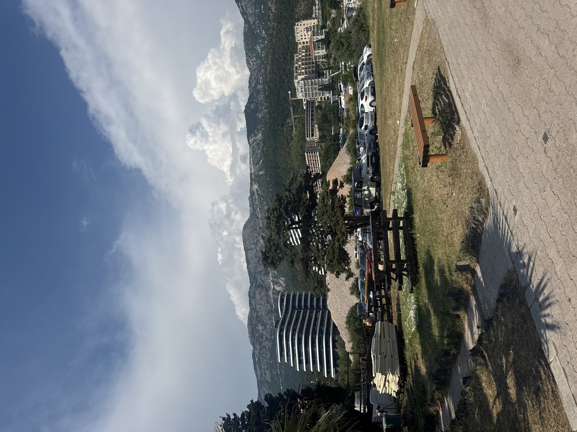 The image depicts a scenic view of Boreti, Montenegro. In the foreground, a paved walkway with a wooden bench runs alongside a grassy area. Beyond the grass, a row of parked cars is visible, suggesting a public space or parking lot.
The middle ground is dominated by a striking, modern building with a curved, stepped facade, set against a backdrop of lush green mountains. Further up the mountainside, several other buildings, likely hotels or residential complexes, are nestled amongst the trees. A construction crane is also visible, indicating ongoing development.
The background features a vast expanse of blue sky filled with dramatic white clouds. The mountains rise steeply, covered in dense vegetation. The scene appears to be captured during daylight, with the sun casting distinct shadows on the walkway, suggesting it is likely afternoon. The overall atmosphere is one of a tranquil coastal resort town with a mix of natural beauty and modern infrastructure. There is no visible text in the image.