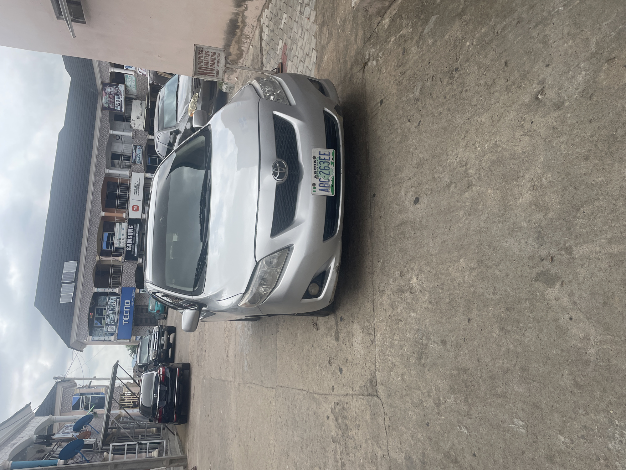 A silver Toyota Corolla is parked on a concrete road in front of a commercial building. The car has a license plate that reads "ABUJA ABC-2633EE". The building behind the car has a dark roof and a facade with several shops. Signs on the building advertise "TECNO", "SAMSUNG", and "EMI'S UNISEX SALON". There are other cars parked in the background, including a black sedan and a dark-colored SUV. The sky is overcast. There is a "NO PARKING WAITING HAWKING KEEP MOVING" sign visible to the right of the car. This scene is likely located in Jalingo, Nigeria.