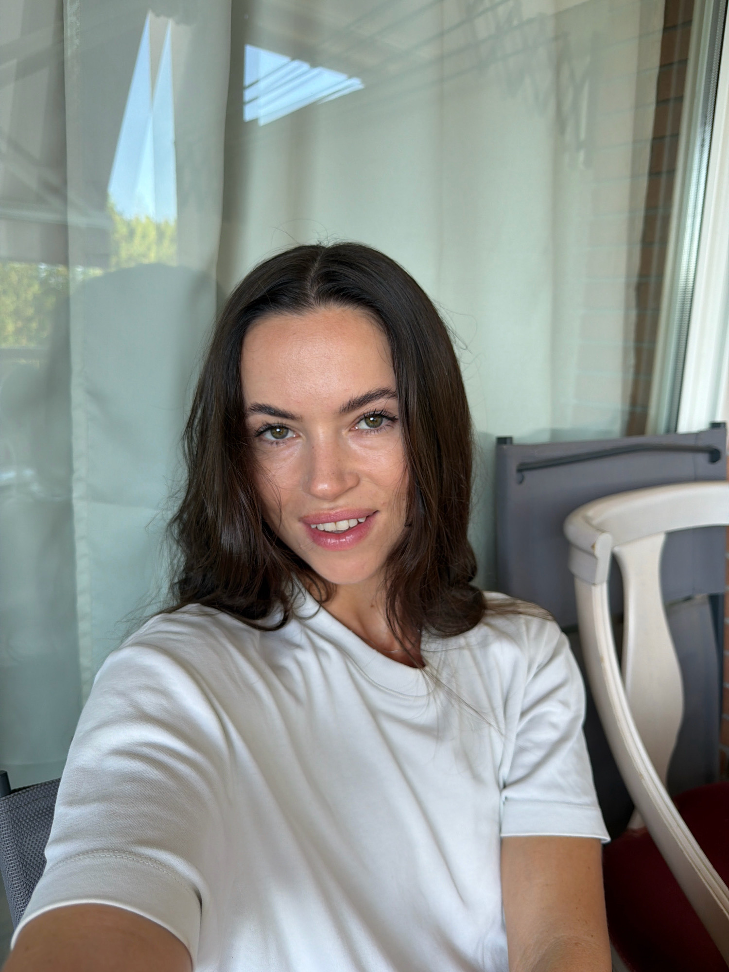 A young woman with long, dark brown hair is taking a selfie in what appears to be an outdoor seating area. She is wearing a plain white t-shirt. Her expression is neutral with a slight smile. Her eyes are open and looking directly at the camera. The background shows sheer white curtains and a window with reflections of foliage and architectural elements. Behind her, a grey chair and a portion of a white-painted chair are visible. The lighting suggests it is daytime. The setting is likely a balcony or patio in a residential or hospitality building.