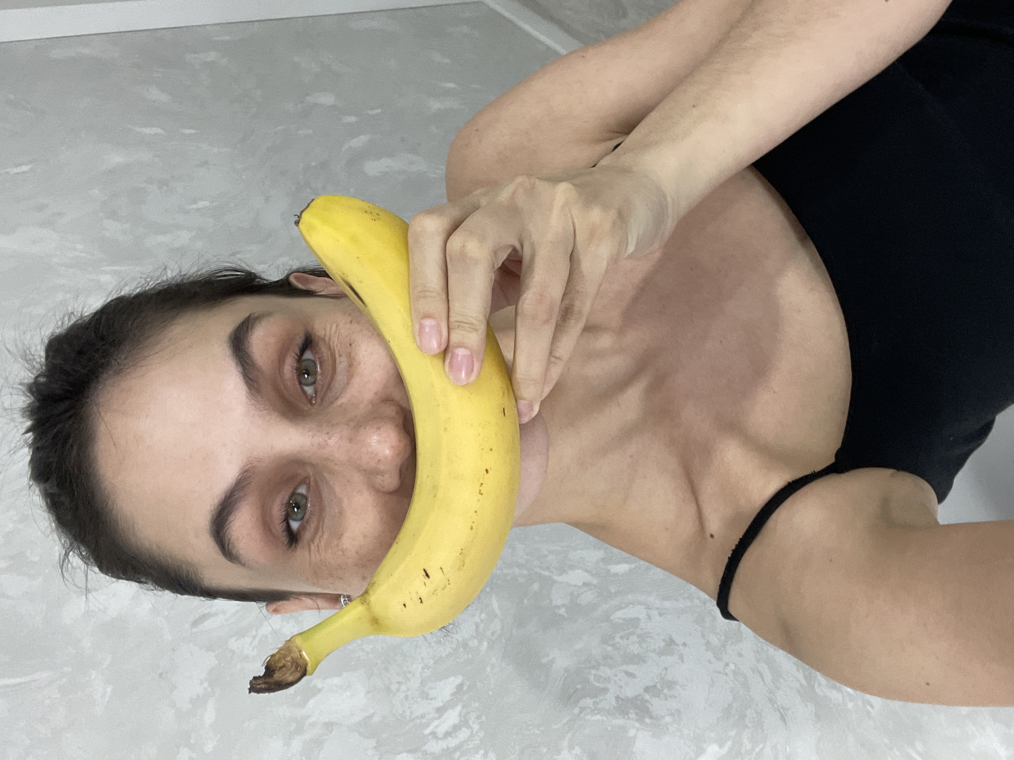 This image, taken indoors in Izhevsk, Russia, features a person, likely female, playfully holding a ripe yellow banana. The photograph is oriented sideways, with the subject's head positioned on the left side of the frame and her body extending towards the right.
The central activity involves the person holding the banana horizontally across the lower half of her face, giving the impression of a banana-shaped smile. Her eyes, which appear to be a greenish-hazel color, are open and looking directly at the camera. Freckles are visible on her face, and her eyebrows are well-defined. One hand is visible holding the banana, which shows some small brown spots, indicating its ripeness. Part of her arm and shoulder are also visible, and she is wearing a black top or strap.
The background is a light gray, textured surface, suggesting an indoor wall or floor. The lighting is bright and even, illuminating the subject clearly. The overall impression is one of playfulness and lightheartedness. There are no visible texts or specific location cues beyond the provided context of Izhevsk, Russia. The time of day and weather are indeterminate due to the indoor setting.