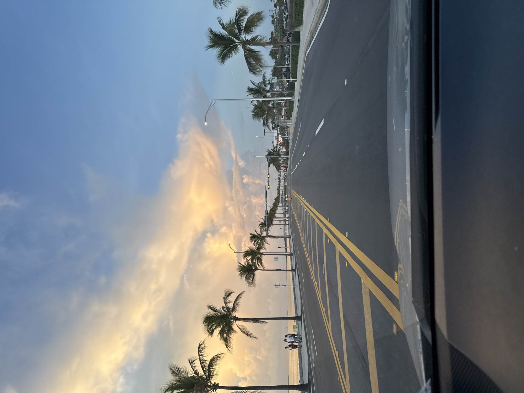 The image depicts a coastal highway with palm trees lining the left side, leading towards the ocean. The sky is a mix of blue and dramatic clouds tinged with sunset colors. Several people are visible walking on a sidewalk adjacent to the ocean. The road is paved and has yellow double lines indicating traffic lanes, with white lane markers. Streetlights are visible along the road. In the distance, there are buildings and more palm trees. The foreground shows the dashboard and windshield of a vehicle. The scene is captured during what appears to be either sunrise or sunset due to the lighting and cloud colors. The location is the Overseas Highway (US Highway 1) in Key West, Florida, USA, a scenic route known for its ocean views and palm-lined streets.