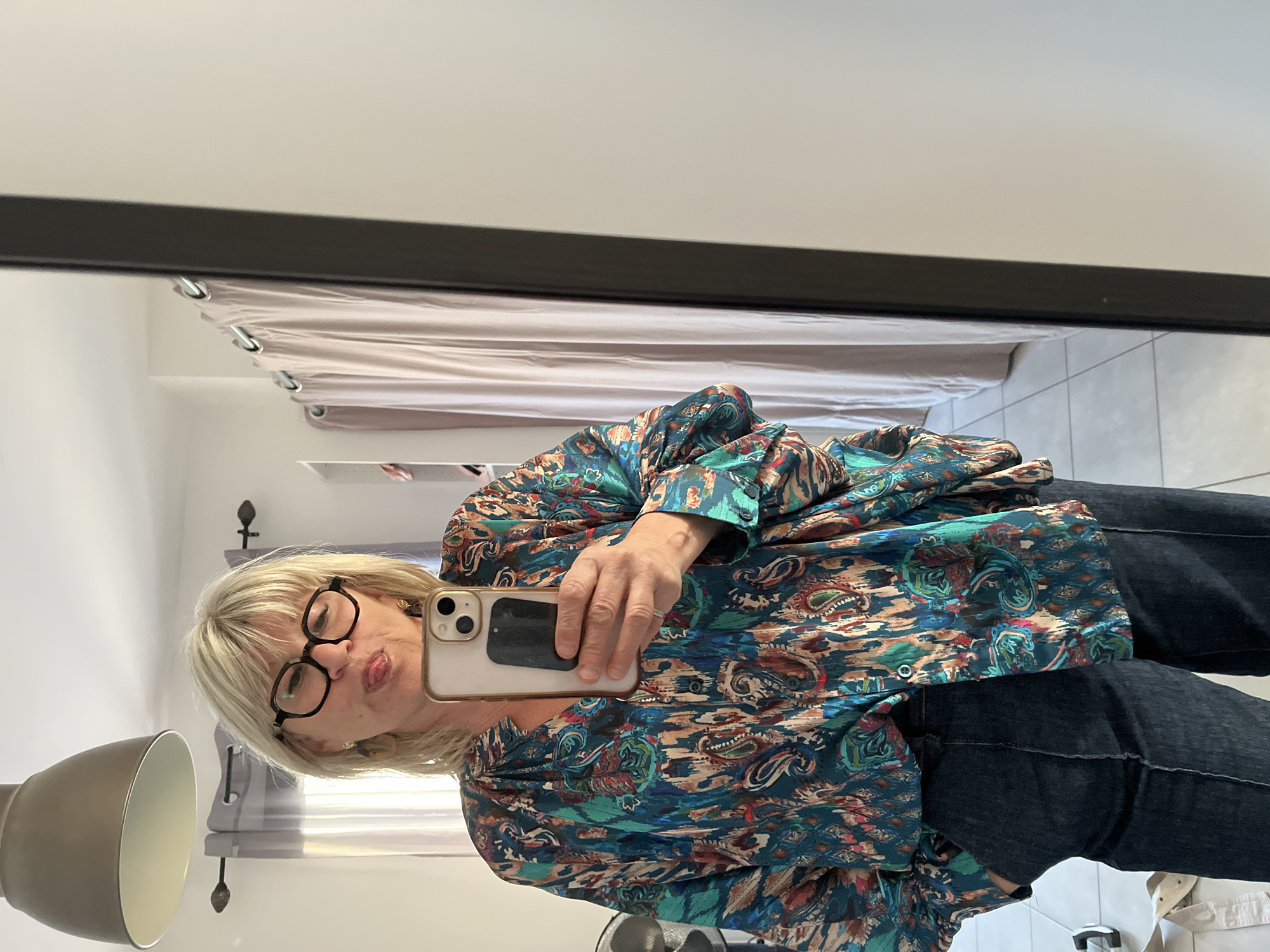 A fair-skinned woman with blonde hair and glasses poses for a selfie in a mirror. She is wearing a patterned shirt and jeans. Behind her, curtains and a lamp are visible. The location is likely indoors in Chambœuf, France.