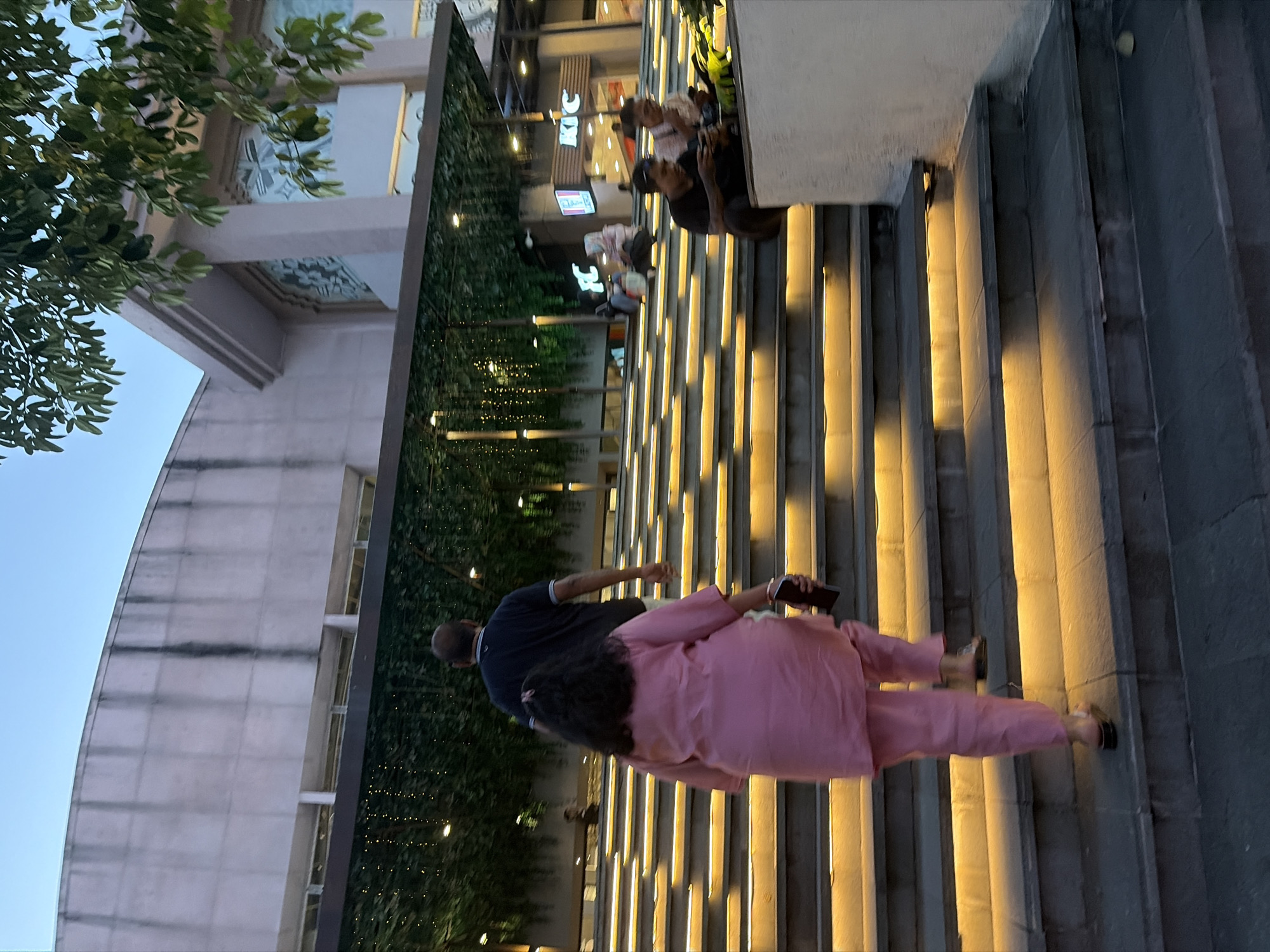 The image shows an outdoor view of a modern shopping mall in Kuta, Indonesia, at dusk. A man in a dark blue t-shirt and a woman in a pink salwar kameez are walking up a wide, well-lit staircase. The steps are illuminated with warm yellow lights. The building has a light-colored facade with large windows. Green foliage with hanging lights is visible above the staircase. In the background, there are signs for KFC. Other people are seated on the stairs in the distance, some looking at their phones. The sky is a soft blue, indicating the transition from day to night.