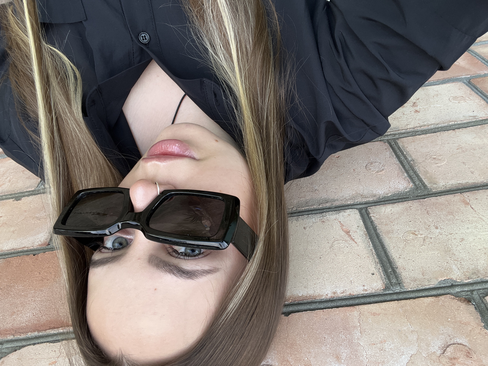 An overhead shot depicts a young woman posing, appearing upside down against a backdrop of reddish-brown brickwork, likely a paved ground or wall in Novopokrovskaya, Russia.
The primary subject is a young woman with long, straight hair, which is predominantly brown with distinct blonde highlights fanning out around her head. She is wearing a black button-up shirt, partially unbuttoned, revealing a glimpse of her chest and a thin black string necklace. Her face is central to the image, featuring glossy pink lips and a small silver hoop nose ring on her left nostril. She is wearing stylish, square-framed black sunglasses, beneath which her blue eyes are visible, looking directly towards the camera. Her eyebrows are well-defined, and mascara highlights her eyelashes.
The activity appears to be posing for a photograph. The perspective is from above, emphasizing the woman's face, hair, and upper body in relation to the textured brick surface. The natural lighting suggests it is daytime. There is no visible text in the image.