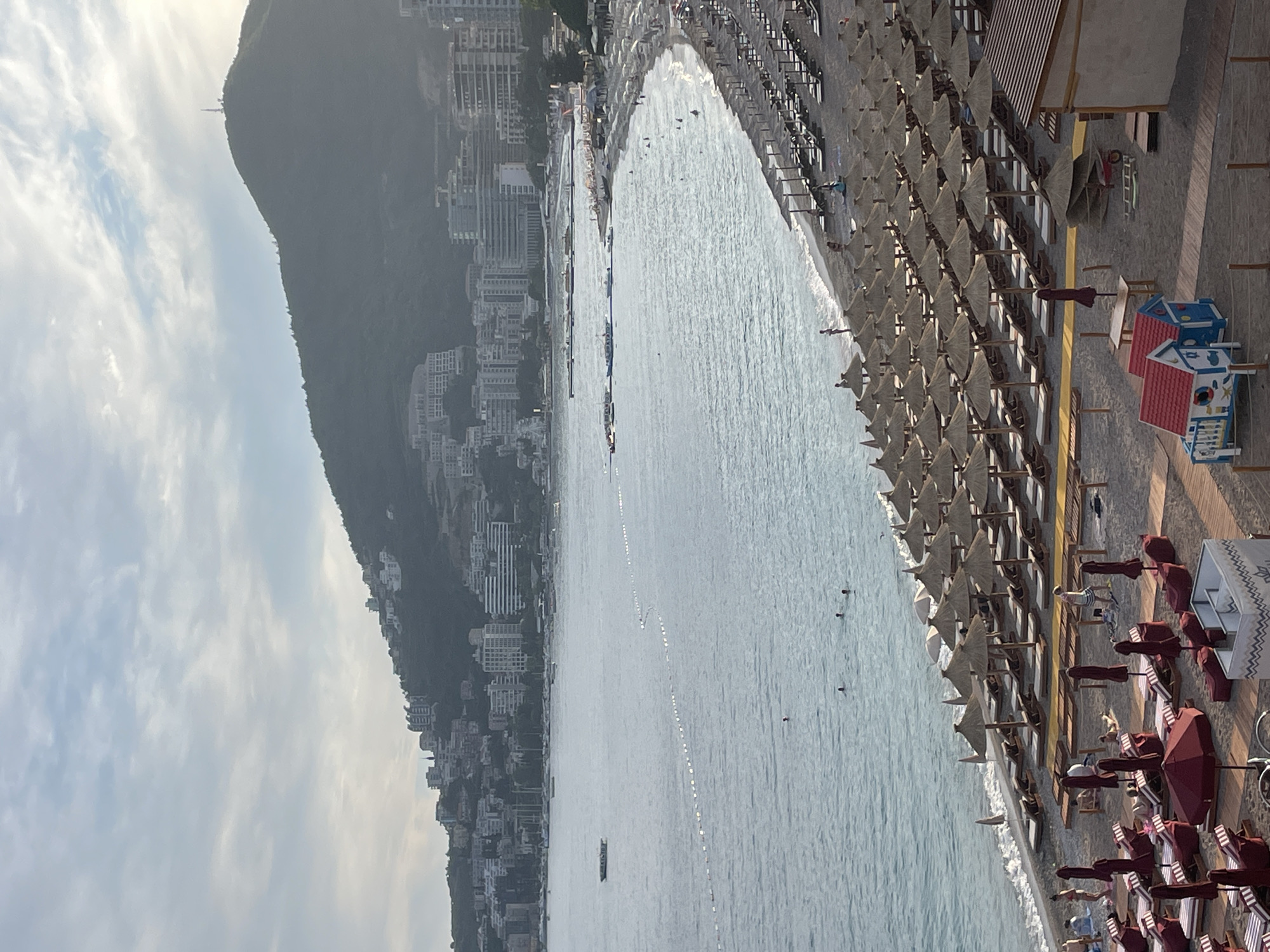 The image captures a coastal scene in Budva, Montenegro, likely during the daytime, with overcast skies suggesting a diffused light. A crescent-shaped beach is densely populated with rows of lounge chairs and straw-umbrellas, indicating a well-established tourist resort. The beach sand is dark and appears to be packed. The calm, light-blue water of the Adriatic Sea gently laps at the shore.
In the sea, several small boats are visible, along with a demarcation line of buoys stretching across the water. A few individuals are swimming in the sea. Beyond the beach, a town sprawls up the side of a large, verdant hill. The town is characterized by numerous multi-story buildings, primarily white and light-colored, suggesting hotels and apartments. The architecture is modern and densely packed. A prominent television tower stands atop the highest point of the hill.
The foreground of the image reveals more of the beach infrastructure, including additional seating arrangements and some wooden walkways. A small, brightly colored playhouse for children, resembling a miniature house with a red roof, is situated on the sand. The overall impression is one of a bustling seaside resort area, with a clear juxtaposition of natural landscape and urban development.