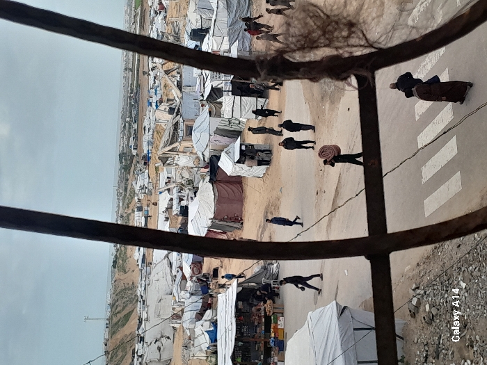 An elevated view depicts a densely populated informal settlement in Khan Yunis, Palestinian Territory, partially framed by dark, rusty metal bars in the foreground. A paved road with a white pedestrian crossing is visible in the lower center, where two individuals in dark clothing, one wearing a brown robe, walk from right to left.
In the midground, a dirt path or open area hosts approximately twenty individuals moving or standing in small groups. One man is observed carrying a rolled, reddish-brown mat on his left shoulder, walking leftward. To the left of the path, a makeshift market stall with a white canopy displays goods. The settlement spans across the midground and background, characterized by numerous white and beige tents, tarpaulins, and rudimentary structures constructed from various materials, many appearing damaged or partially collapsed.
Beyond the encampment, sandy, undulating terrain is visible, topped by a distant communication tower. A hazy urban or semi-urban skyline extends to the horizon. The sky is overcast. The image bears a "Galaxy A14" watermark in the lower left corner.