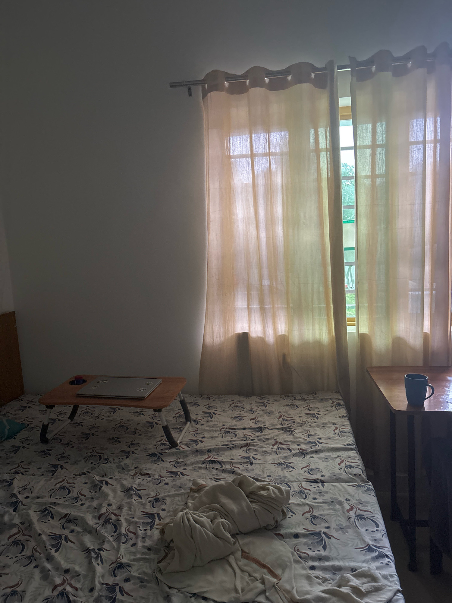 The image depicts a dimly lit bedroom in Bhatta, India. A bed with a floral patterned bedsheet dominates the foreground, partially covered by a light-colored blanket. On the bed, a portable laptop table holds a laptop. To the right of the bed, a small wooden table has a blue mug on it. The room is illuminated by natural light filtering through sheer curtains covering a window. The overall scene suggests a lived-in, personal space.