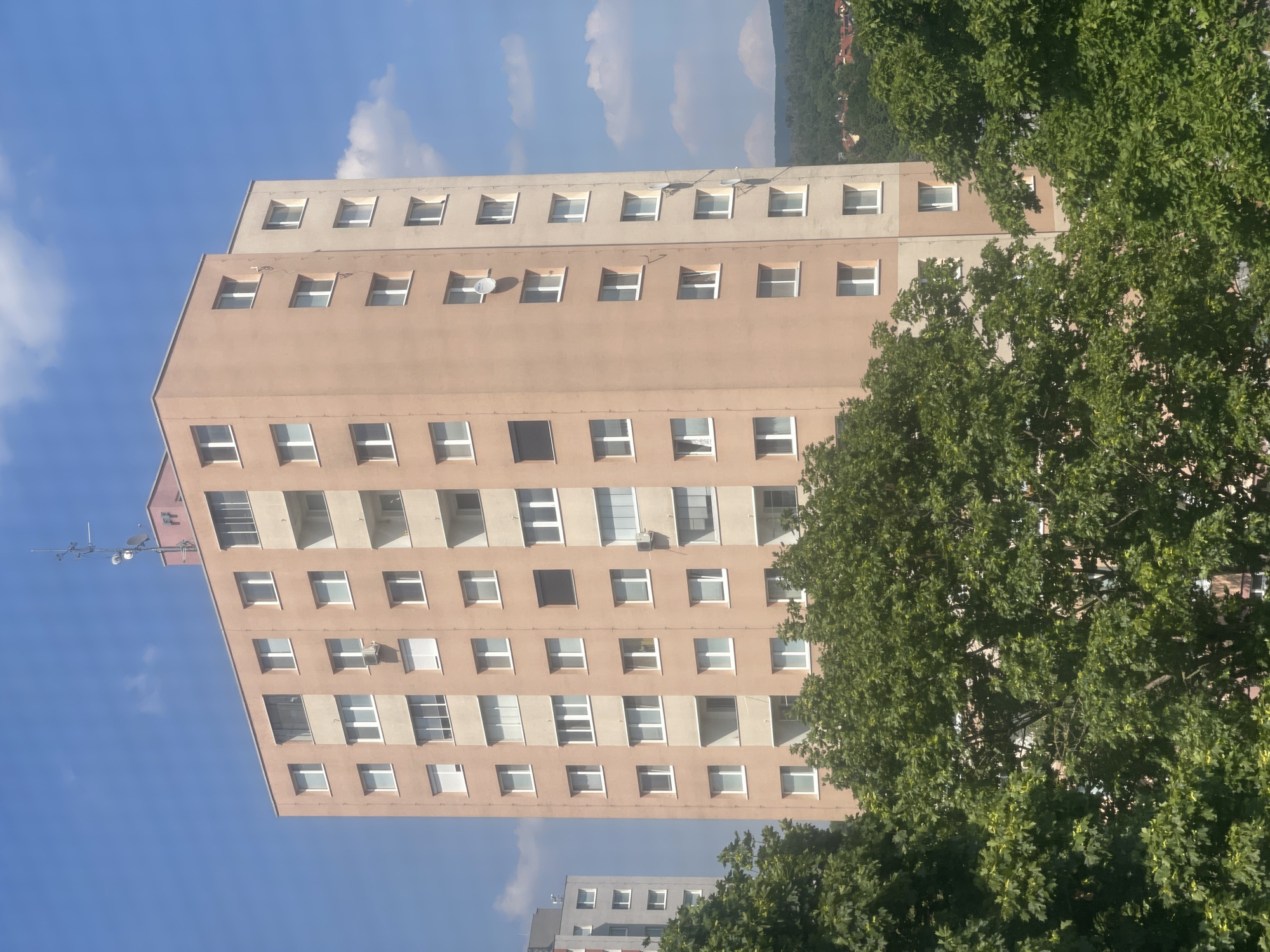 The image captures a tall, multi-story apartment building in a city, with a clear blue sky and scattered clouds overhead. The building is light brown or peach-colored with rows of uniformly sized windows and balconies on each level. A smaller, gray apartment building is partially visible to the left. Lush green trees in the foreground partially obscure the lower floors of the main building, suggesting a residential area with green spaces. The background reveals a distant, tree-covered hillside with some buildings, indicating a suburban or outer-city location. There are no people visible in the image. The scene is static and appears to be a daytime exterior shot, possibly taken from a higher vantage point.