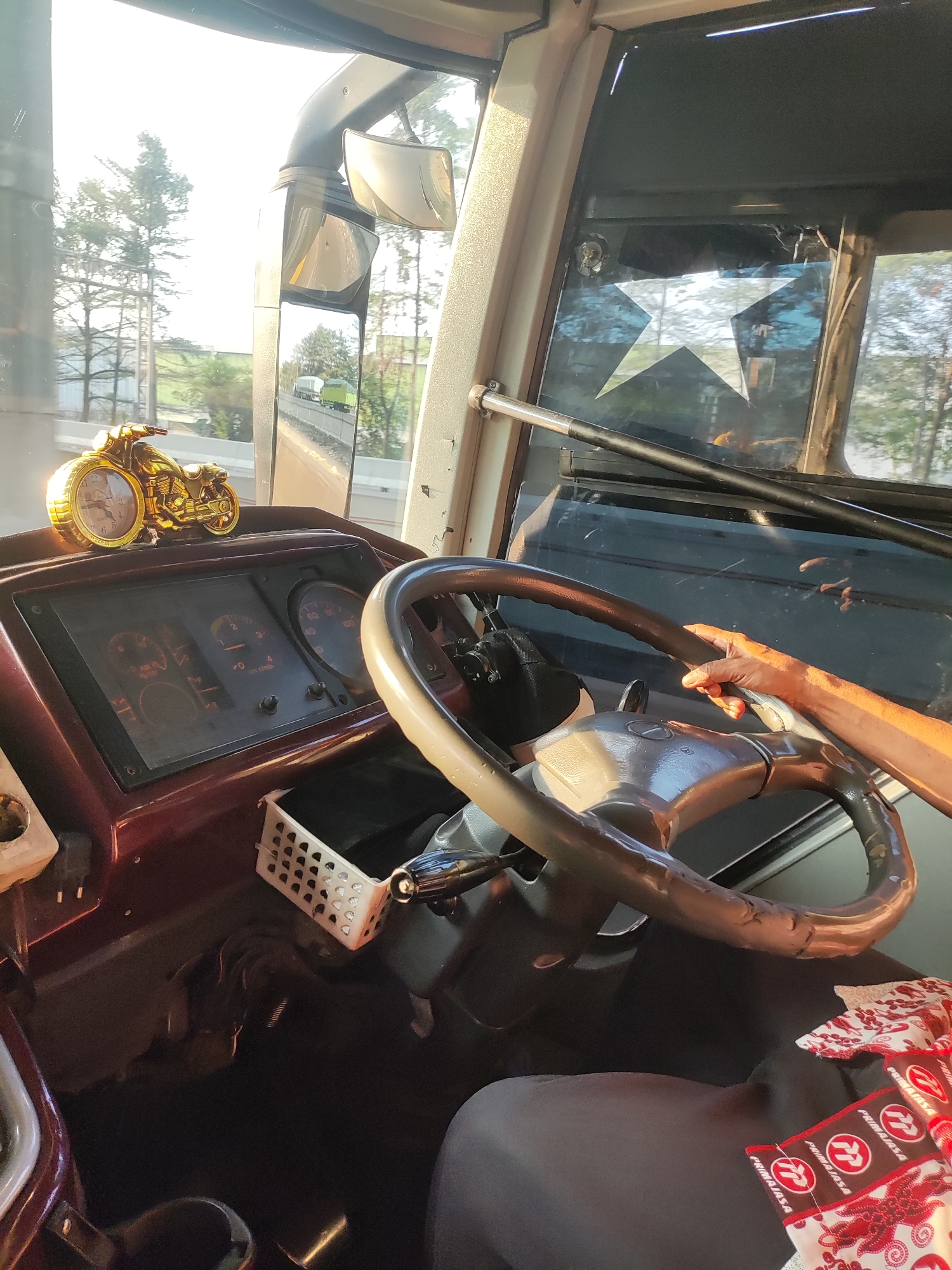 The image shows the interior of a bus's driver's cabin, with a focus on the dashboard and steering wheel. A gold-colored miniature motorcycle clock is visible on the dashboard. The steering wheel is partially obscured by a person's hand, suggesting the bus is in motion. Reflections in the windshield and mirrors indicate an outdoor environment with trees and a road. There are also some decorative elements on the dashboard and window, including a star decal. The overall scene appears to be a snapshot from inside a moving bus, capturing the driver's perspective. The location within Danauindah, Indonesia, is not ascertainable from the visual information provided in the image.