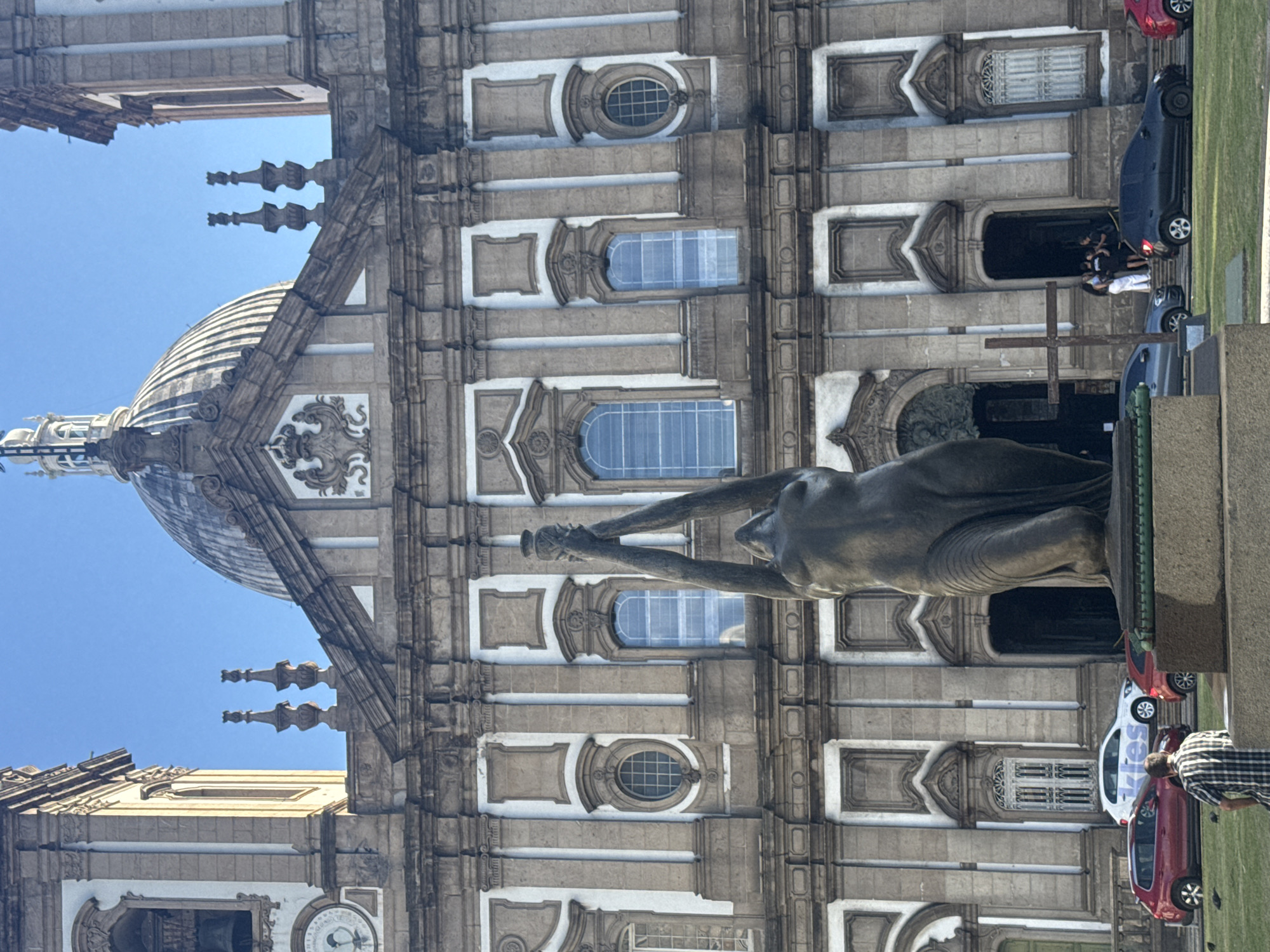 The image shows the Basilica of the Immaculate Conception in Rio de Janeiro, Brazil. A bronze statue of a kneeling figure with arms raised, holding a vase, stands in front of the basilica. The basilica is a large, ornate building with a dome and two bell towers. In the foreground, there is a lawn with several cars parked on it. A man in a plaid shirt is sitting on the lawn, facing away from the camera. There are also a few pigeons on the lawn. The sky is clear and blue.