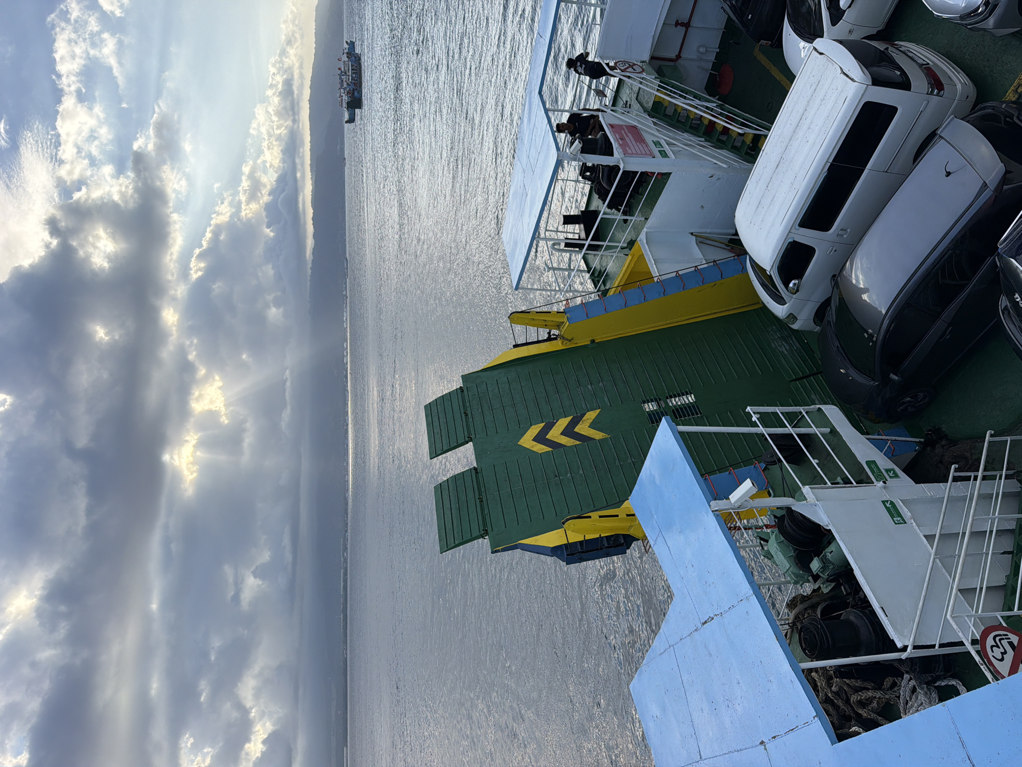The image shows a view from a ferry in the ocean near Melaya, Indonesia. The sky is partly cloudy with sunbeams breaking through, creating a dramatic lighting effect over the water. In the distance, a coastline with hills or mountains is visible. A smaller ferry boat is seen on the water.
The foreground of the image is dominated by the deck of the ferry the photographer is on. There are cars parked on the deck, including a white van and at least two other cars. Ropes and winches are visible on the deck, suggesting the ferry is used for transporting vehicles. A green and yellow ramp or gate is partially raised, indicating it may be preparing to dock or depart. Several people are visible on the ferry's deck, some standing and observing the surroundings. The ferry has blue awnings providing shade. The overall scene depicts a ferry journey in a coastal environment.