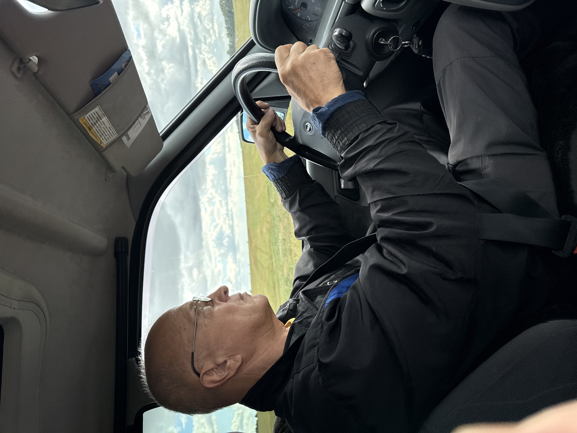 An exterior shot, taken from inside a car, shows a man driving. The man, who appears to be in his late 50s or 60s, has light skin, a bald head, and is wearing glasses, a black jacket, and a yellow shirt. He is holding the steering wheel of the car. The car's interior is visible, including the dashboard, steering wheel, and sun visor. Outside the car, a rural landscape with fields and a cloudy sky is visible. The image does not contain any information about the city of Fominka, Russia.