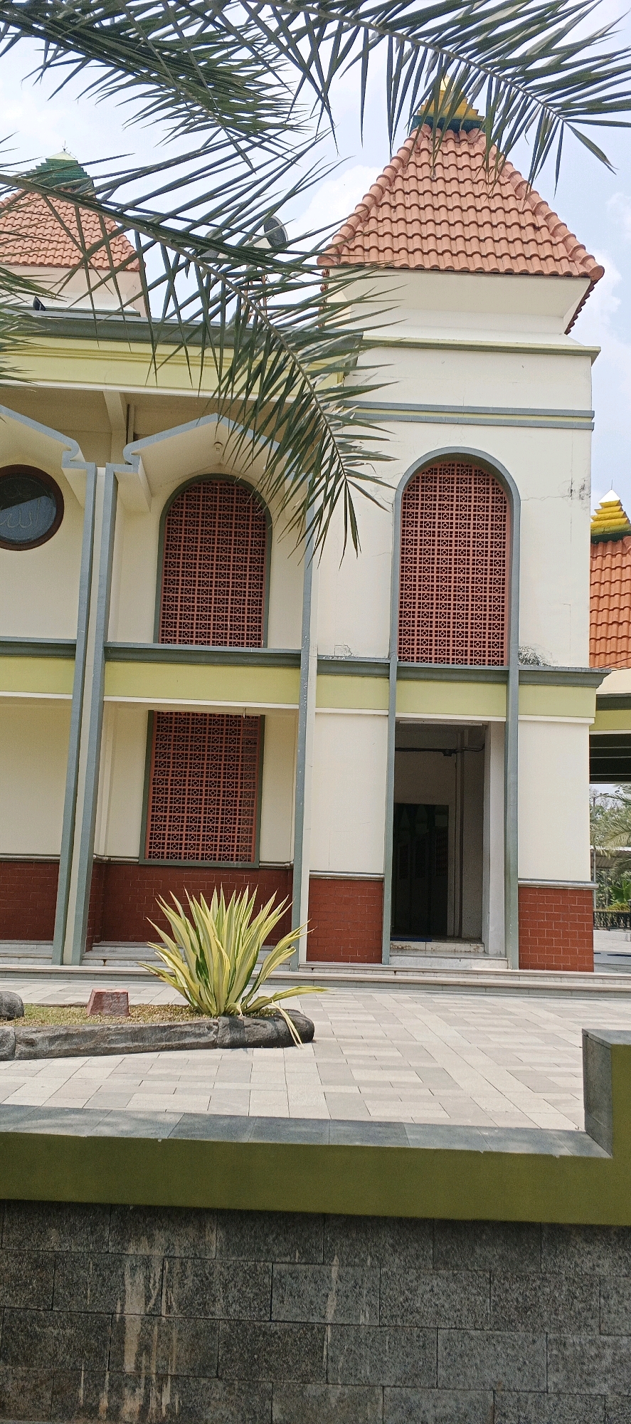 The image shows the exterior of a building with a tiled roof, possibly a mosque, in Kanugrahan, Indonesia. The building has cream-colored walls with decorative red brick accents at the base and arched windows with intricate red latticework. Palm fronds partially obscure the view of the building and the sky, which is visible with some clouds. In the foreground, there is a paved area with a planter containing a spiky green and yellow plant. The ground is paved with rectangular gray stones, and a low wall made of dark, rough-hewn stones is visible at the bottom of the frame. The overall scene depicts an outdoor architectural view with natural elements.